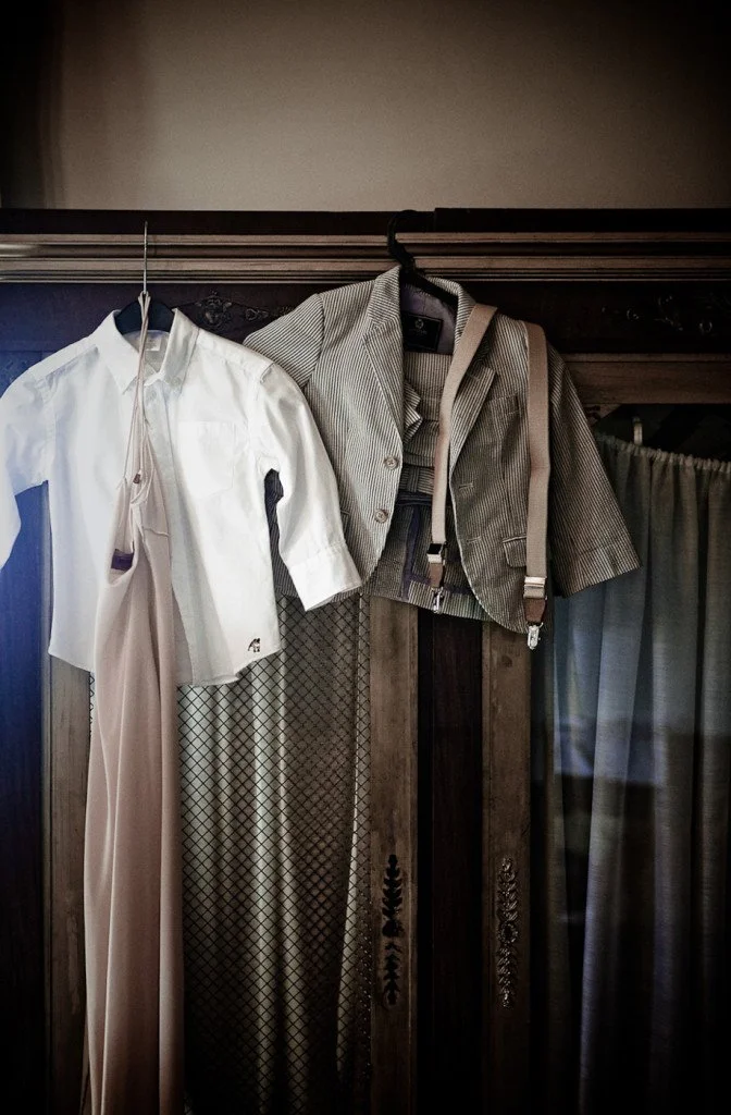 Hanging on a wooden wardrobe are a white shirt on a hanger and a gray plaid blazer with suspenders. A pair of light-colored pants is also hanging underneath the white shirt.