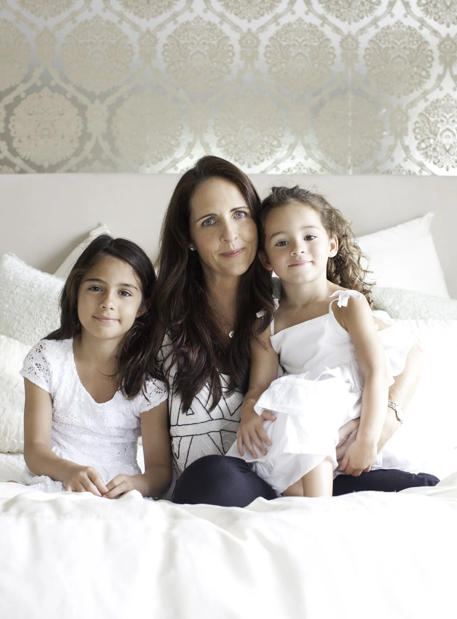 A woman with two young girls sitting on a bed, smiling, with decorative wallpaper in the background.