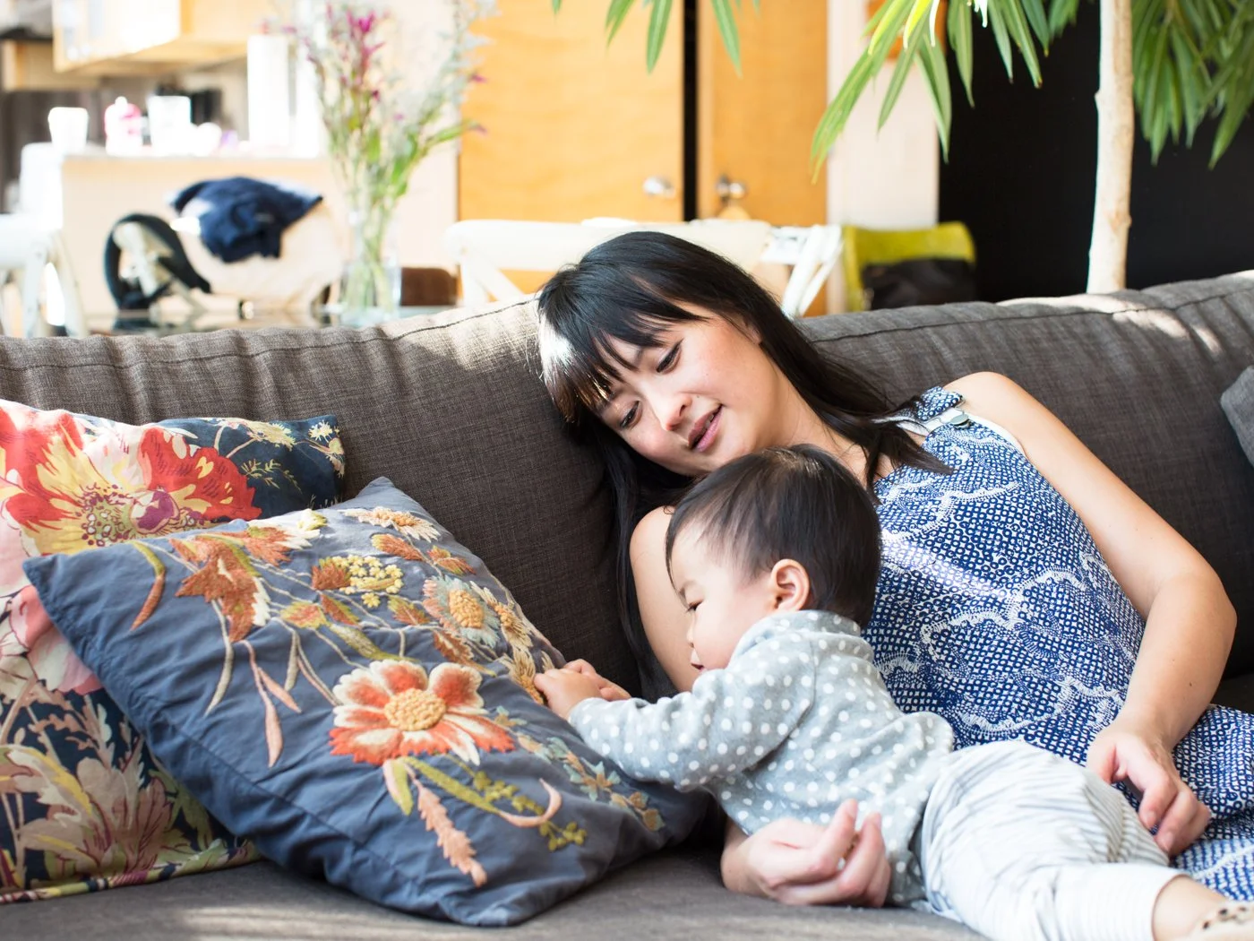 A woman and a young child relaxing on a sofa in a living room, with the woman smiling and looking at the child, who is lying down and reaching towards a pillow with floral patterns.