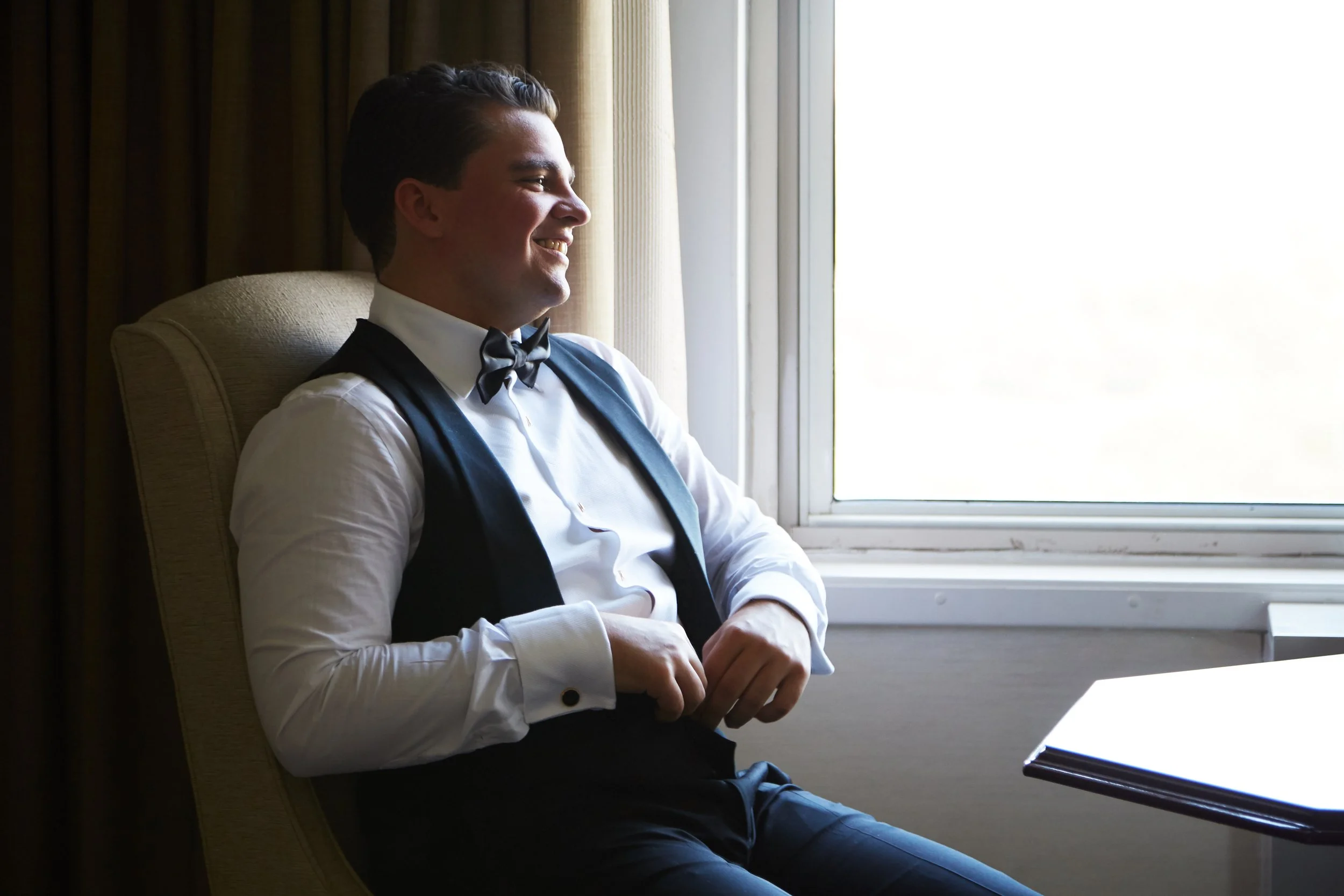 A young man in formal attire, wearing a tuxedo with a bow tie, sitting in a chair by a window with sunlight streaming in.