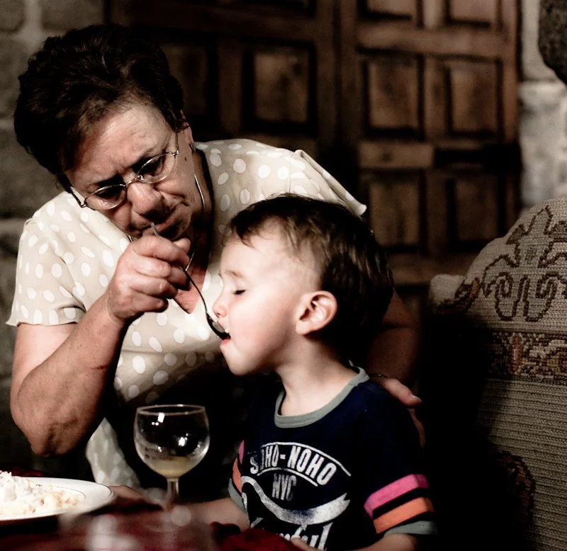 An elderly woman feeding a young boy with a spoon at a dining table in a cozy, rustic setting.