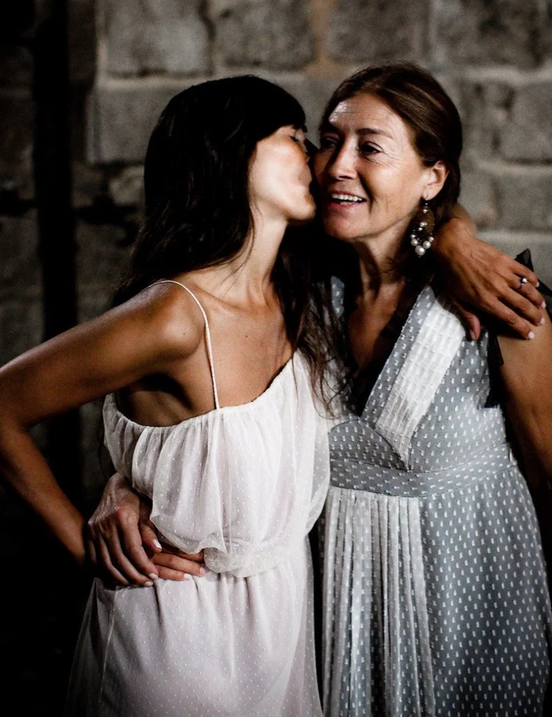 Two women embracing, one is kissing the other on the cheek, both wearing white dresses, against a brick wall background.