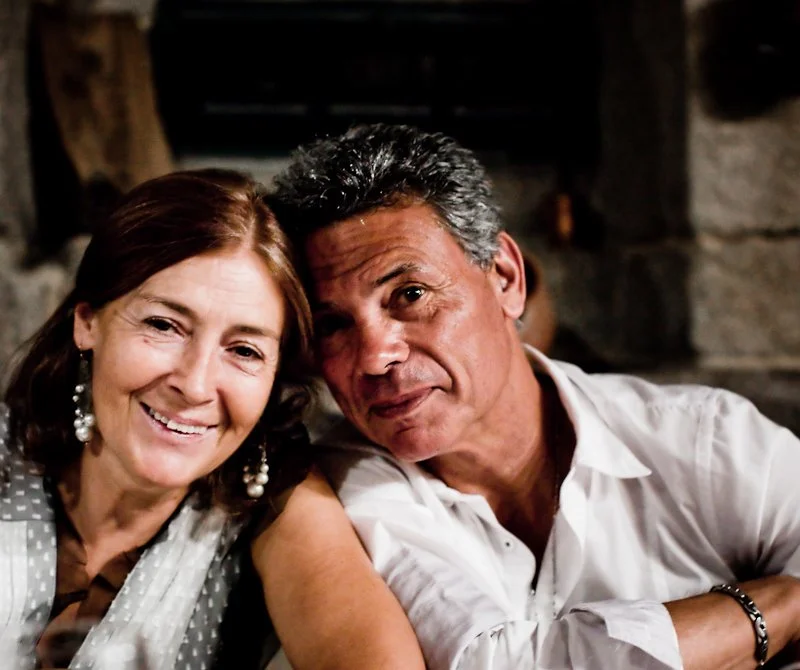Smiling middle-aged woman with earrings and a polka dot scarf posing beside a man against a rustic background