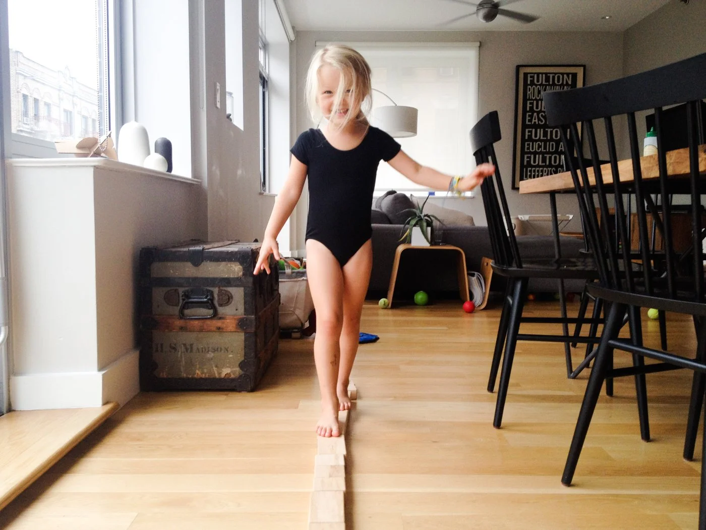 A young girl in a black outfit balancing on a wooden block in a living room with a playful expression.