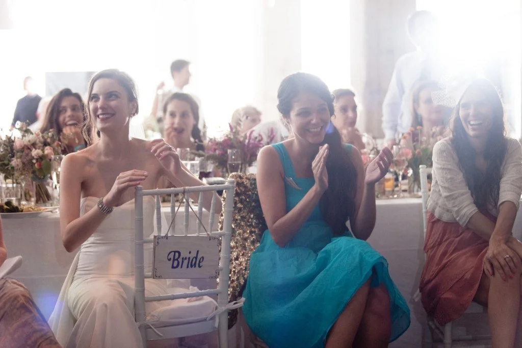 Group of women at a wedding reception, some of whom are sitting at a table, watching and laughing, with a woman labeled 'Bride' in the foreground.