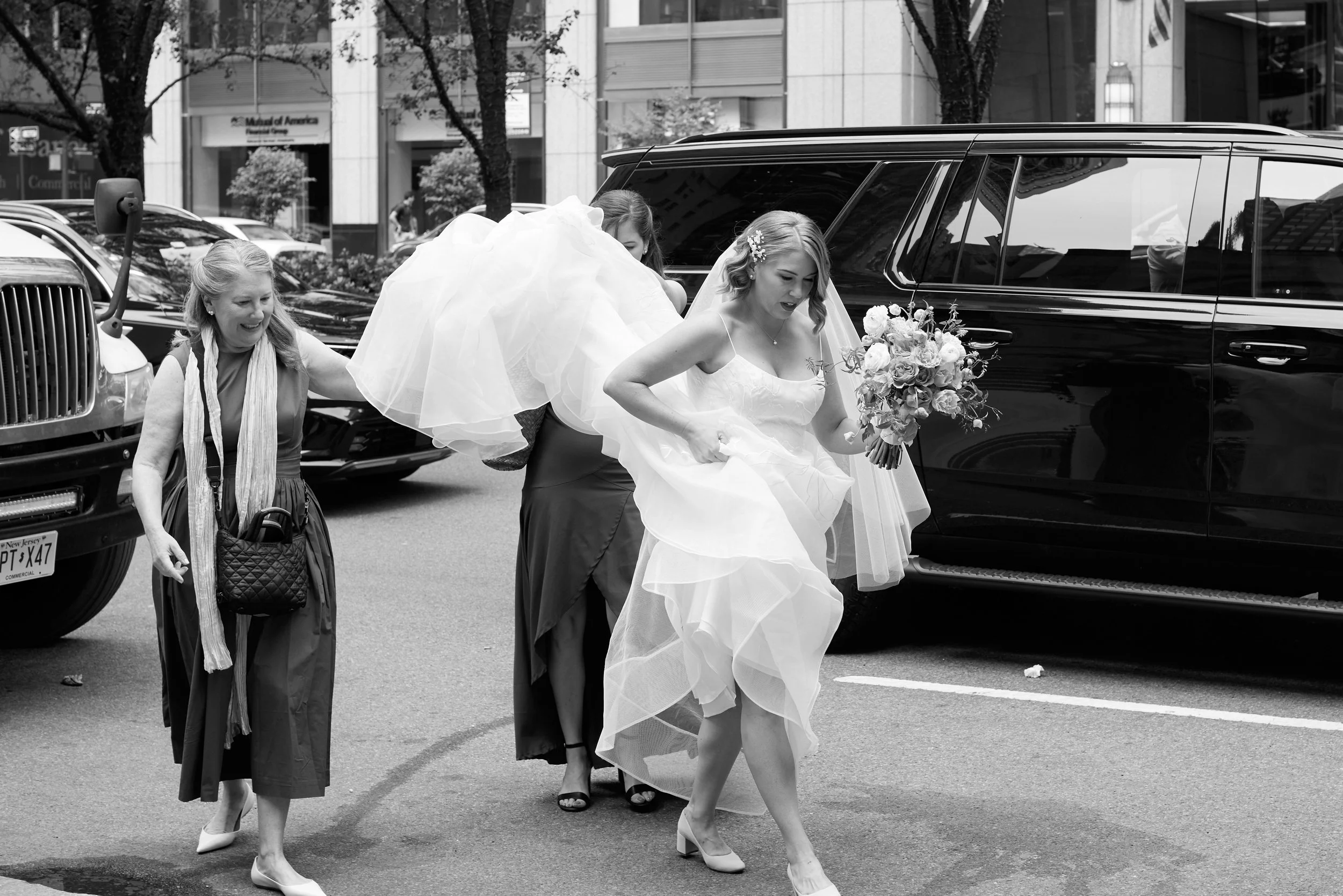 A bride in a wedding dress holding a bouquet of flowers steps over a puddle on a city street, accompanied by her bridesmaids and friends.