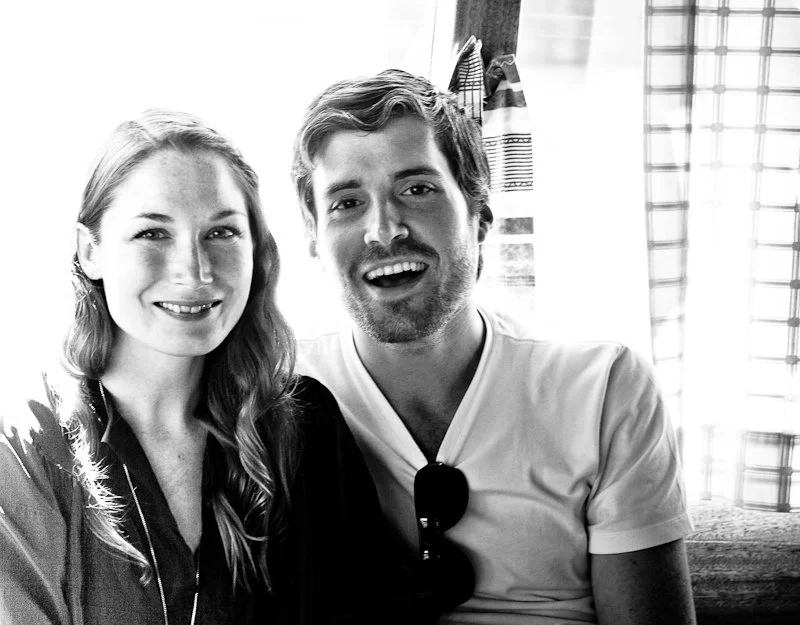 A smiling woman with long wavy hair and a man with short hair and facial hair sitting together indoors, near a window with blinds.