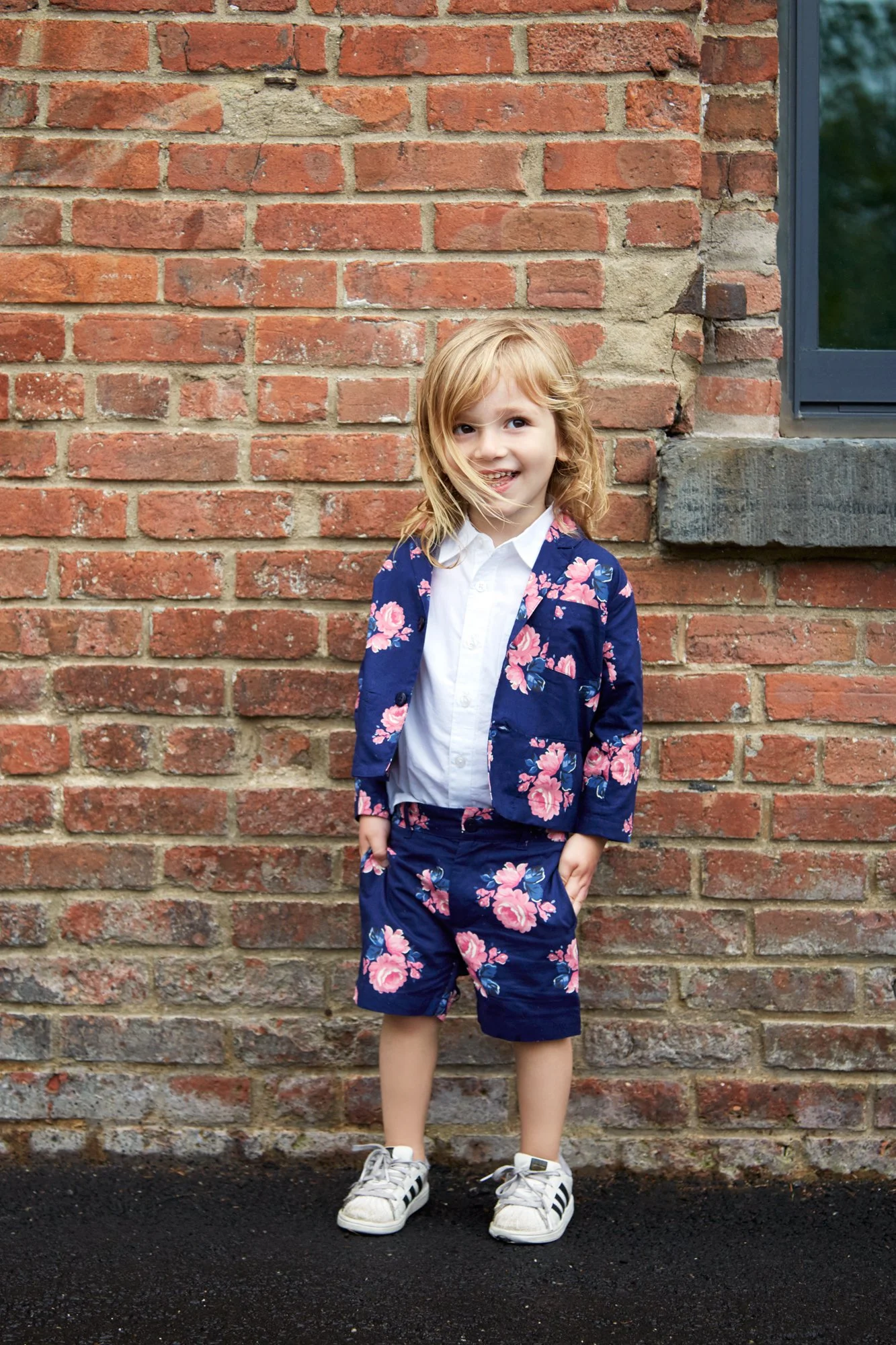 Young girl with blonde hair in a floral blue suit, white shirt, and sneakers standing against a brick wall.