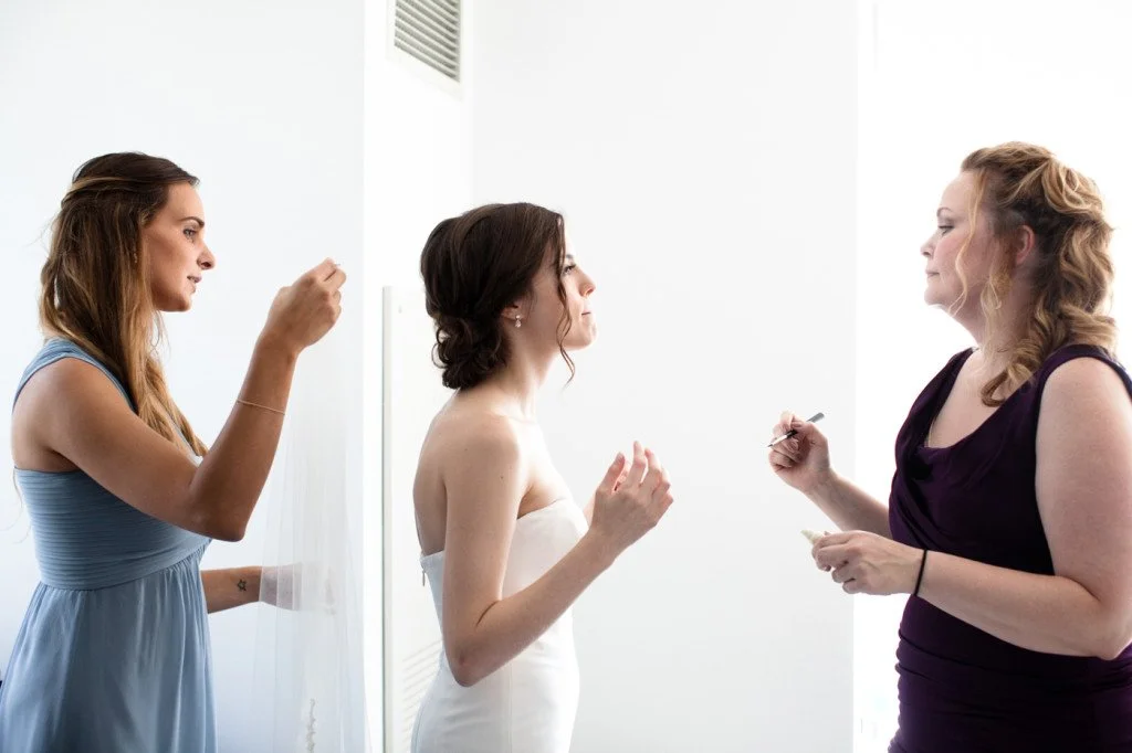 Three women in a room, one in a wedding dress, the other two helping her get ready, with one adjusting her hair and the other applying makeup.