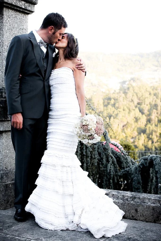 A newlywed couple sharing a kiss outdoors overlooking a scenic landscape. The groom is wearing a black suit and bow tie, and the bride is in a strapless white wedding dress holding a bouquet of flowers.