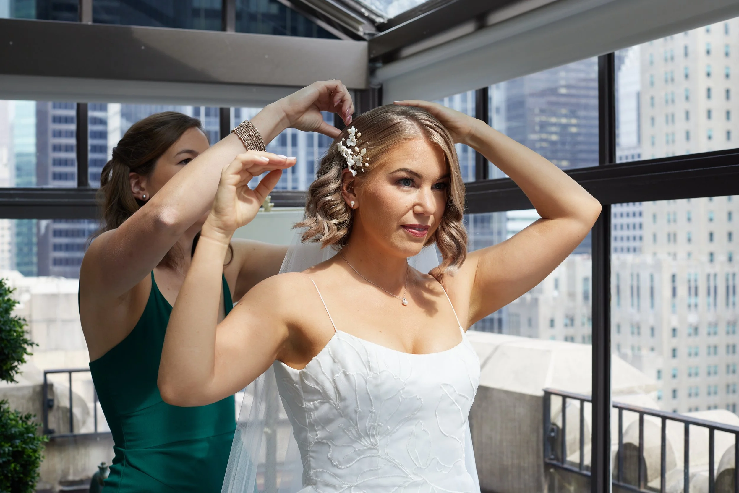 A bride with shoulder-length wavy blonde hair in a white wedding dress adjusting her hair, assisted by a woman in a green dress, in a room with large windows and cityscape views.