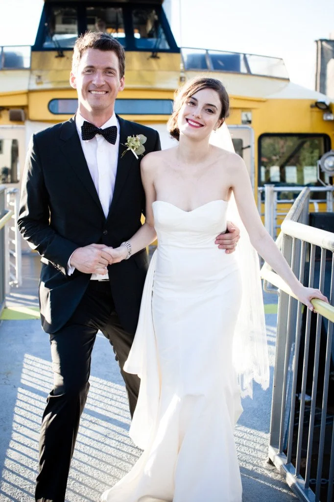 A newly married couple dressed in wedding attire, standing outdoors on a boat deck with a yellow vessel in the background, smiling for a photo.