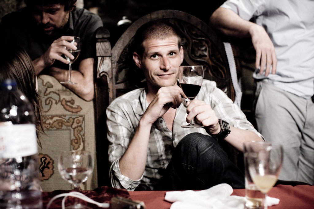A man sitting at a table holding a glass of red wine, looking at the camera, with a slight smile, in a dimly lit setting with other people in the background.