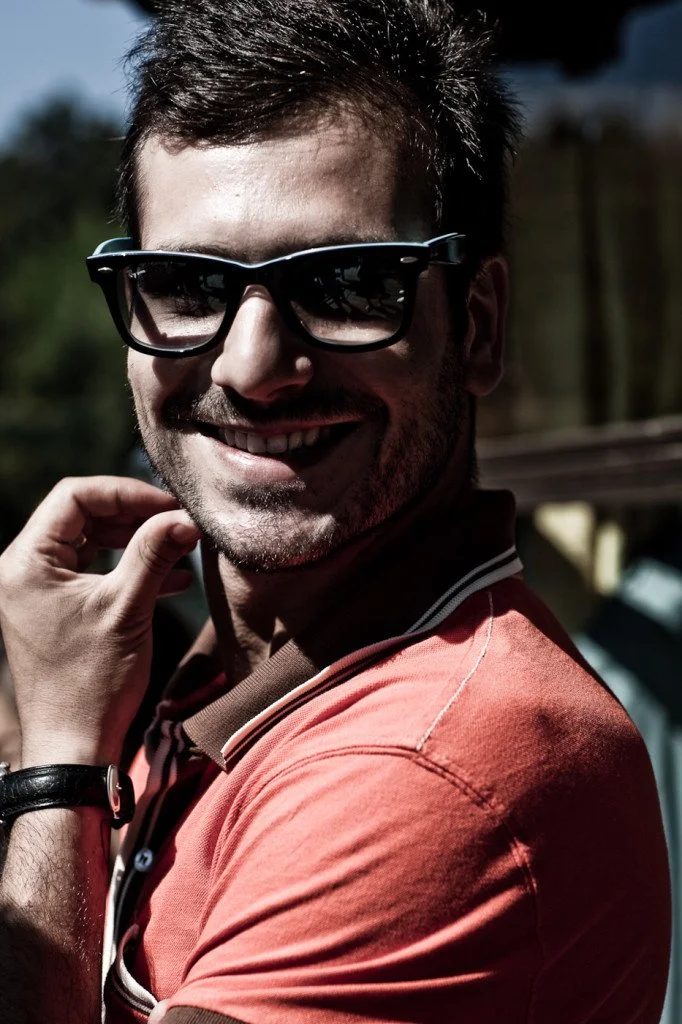 Smiling young man wearing sunglasses and a coral polo shirt outdoors with sunny weather.