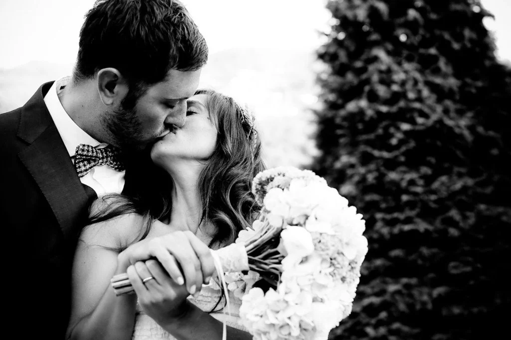 A black and white photo of a newlywed couple sharing a kiss, with the bride holding a bouquet of flowers.