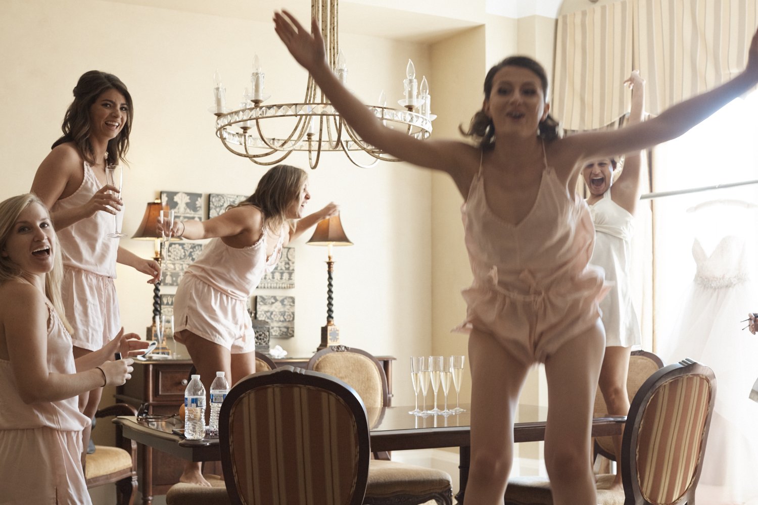 Group of women in pajamas celebrating inside a dining room, some holding champagne glasses, with a woman in the foreground dancing with arms outstretched.