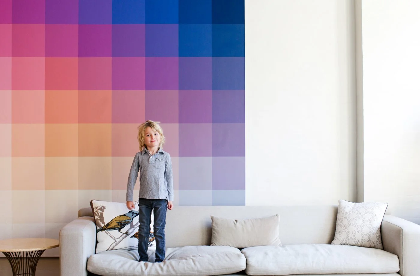 A young girl with blonde hair stands on a cream-colored sofa with multiple pillows, in front of a colorful gradient square wall art, inside a bright room with natural lighting.