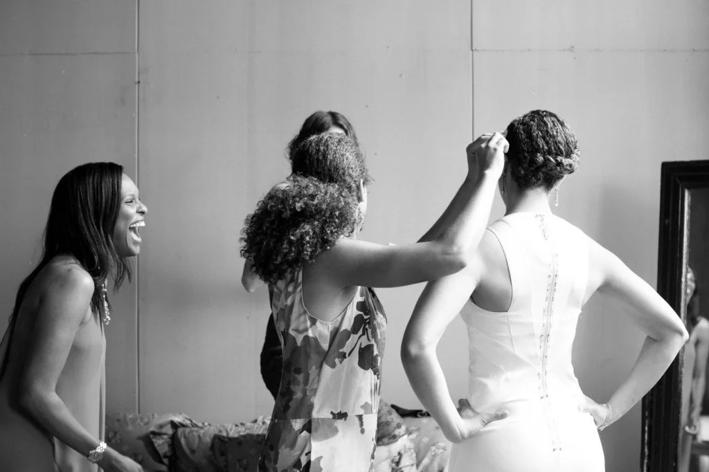 Women dressing and laughing together in a room, with one woman helping another with her hair, in black and white.
