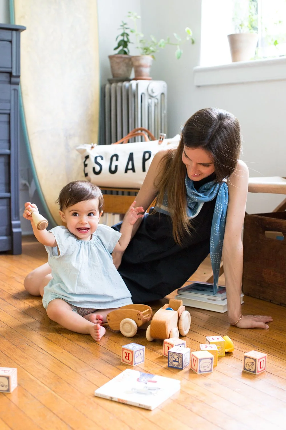 A smiling toddler girl sitting on the wooden floor with toys and blocks, and a woman lying on her side beside her, laughing and engaging with the child, in a cozy room with natural light and plants.