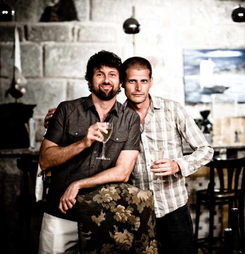 Two men standing together in a cozy, dimly lit bar or restaurant, holding wine glasses and smiling at the camera.