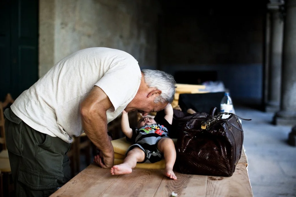 An elderly man leans over a young child lying on a table, looking at the child affectionately in a rustic indoor setting with a large bag and water bottle nearby.