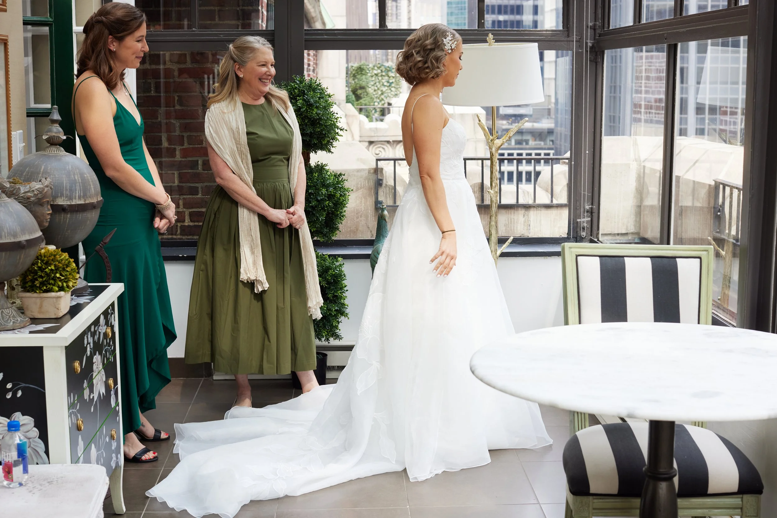 A bride in a white wedding gown standing indoors with three women, one in a green dress and two in casual clothing, smiling and looking at her as she prepares for her wedding.