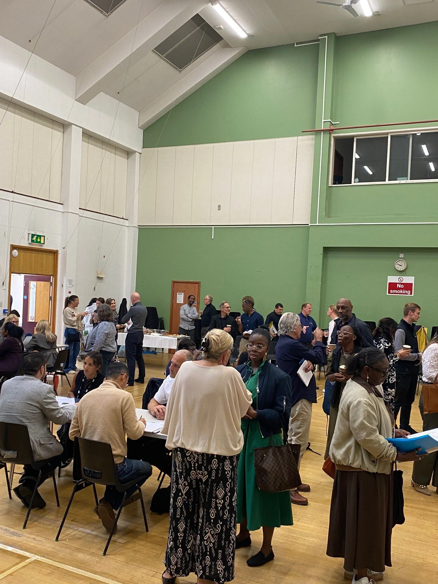 A diverse group of people gathered in a large indoor space with high ceilings, green and white walls, and a wooden floor. Some are standing and talking, while others are seated at tables. There are windows, a clock, a 'No smoking' sign, and exit signs visible in the background.
