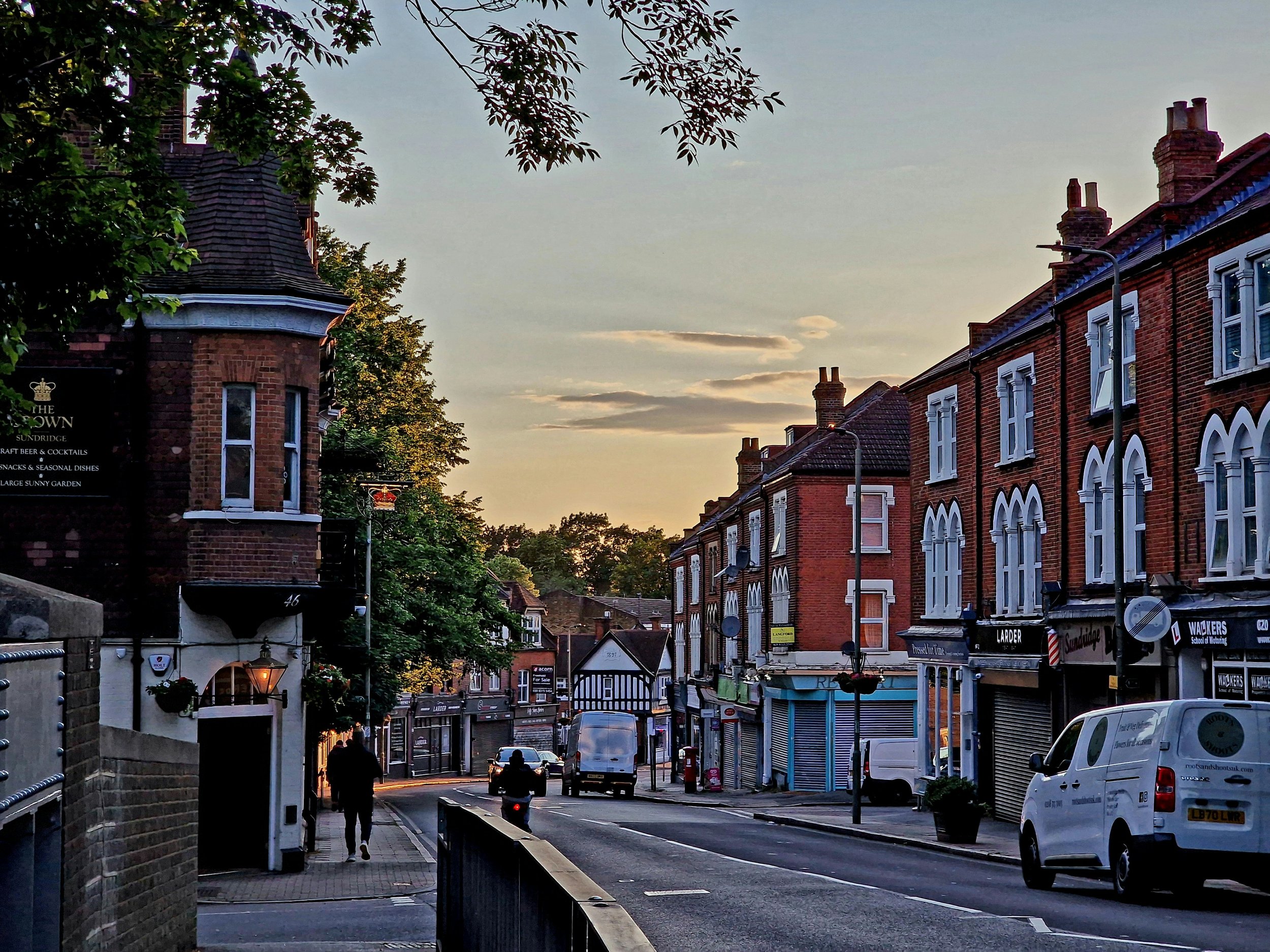 A street scene during sunset with brick buildings, cars parked along the side, and a few people walking. The sky shows soft clouds with a warm glow from the setting sun.