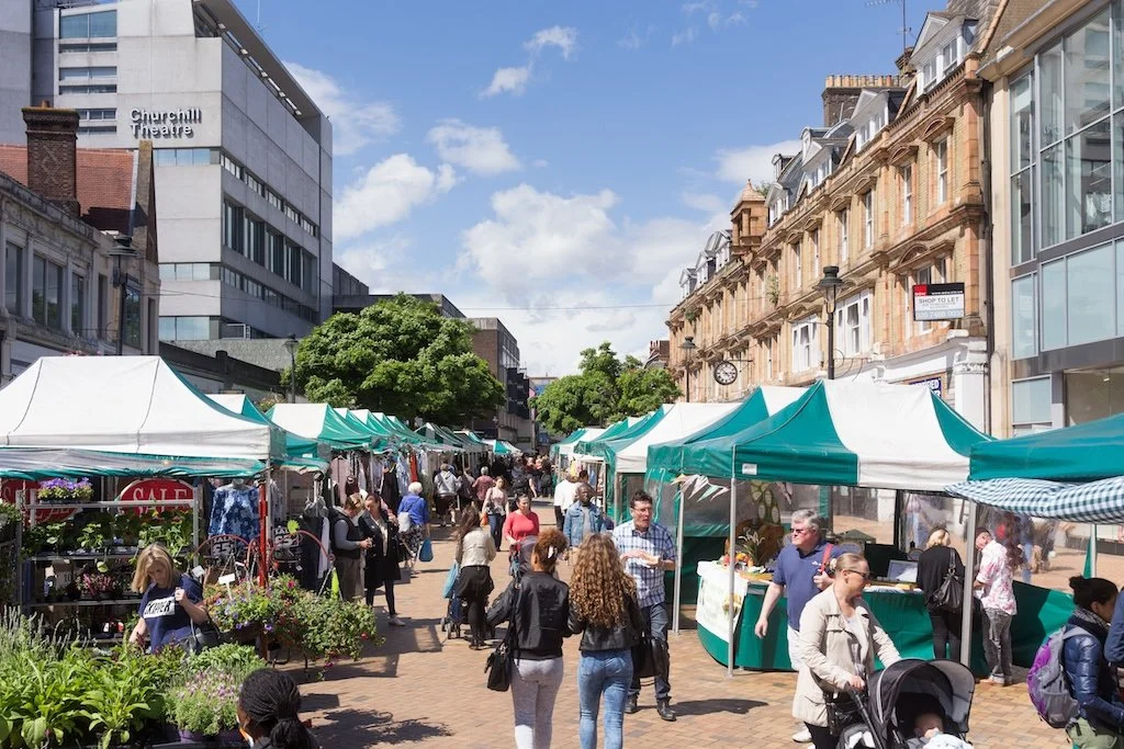 Outdoor street market with various stalls and tents, people browsing, shopping, and walking on a sunny day, with buildings in the background.