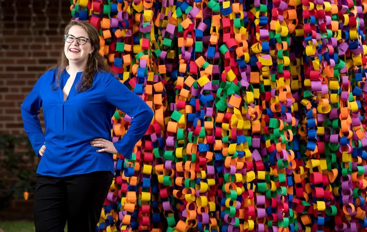 The Wishing Tree's creator, Liz Baker Bowman in a blue blouse, standing outdoors in front of a colorful, spiraled paper chain tree, smiling with hands on her hips.