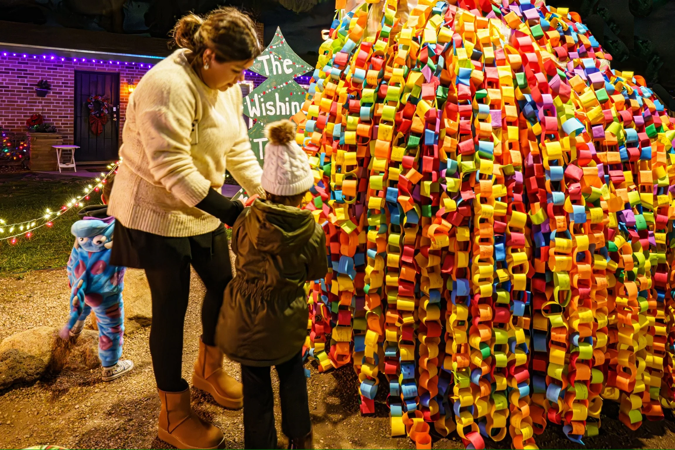 A woman and two children stand in front of Kathleen Bethel's Wishing Tree with colorful paper chains of wishes. The woman appears to be helping the children look at the tree.