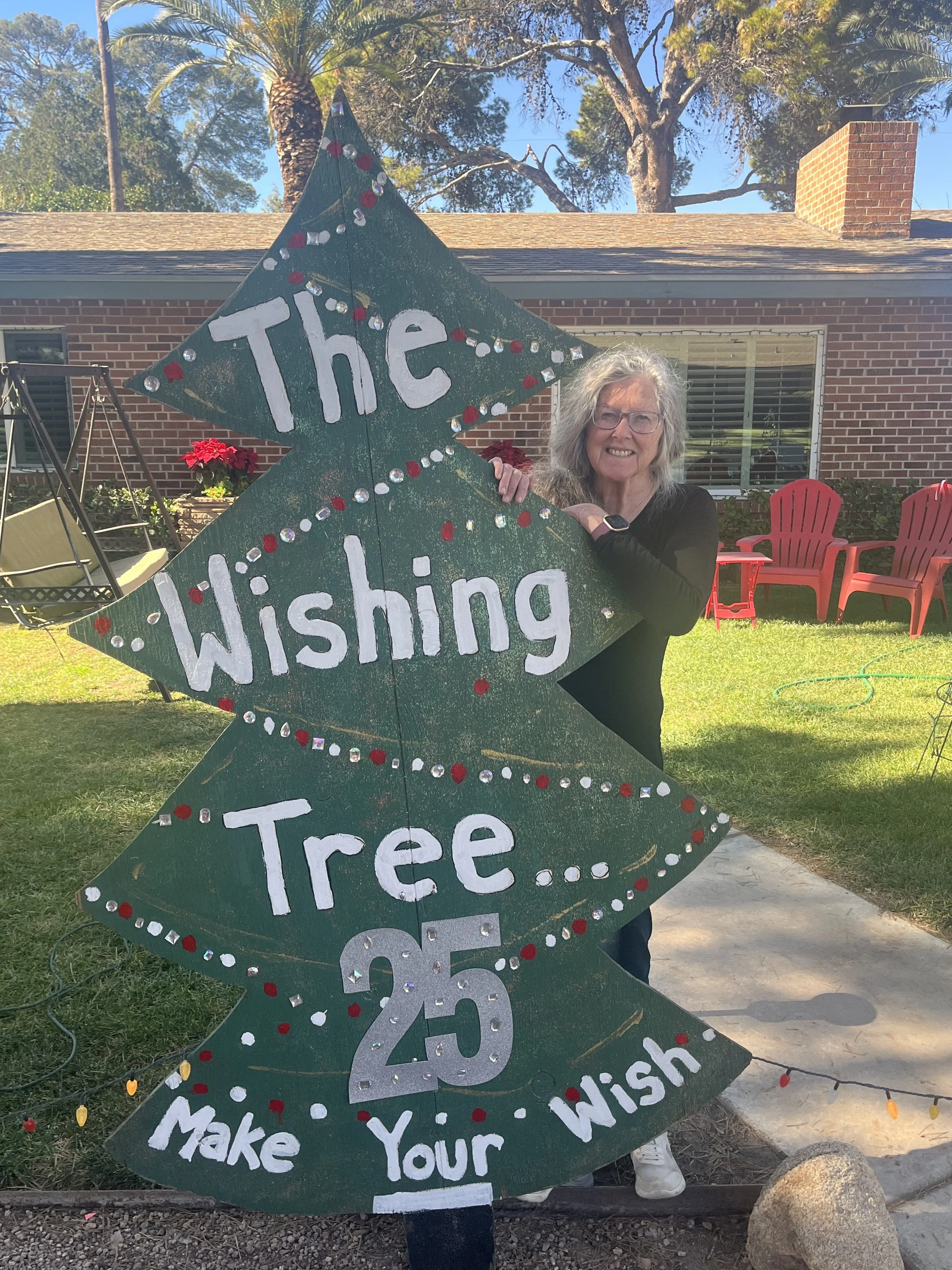 The Wishing Tree's homeowner, holding a decorated sign in the front yard. The sign reads: "The Wishing Tree... 25 Years - Make Your Wish" and is shaped like a Christmas tree.