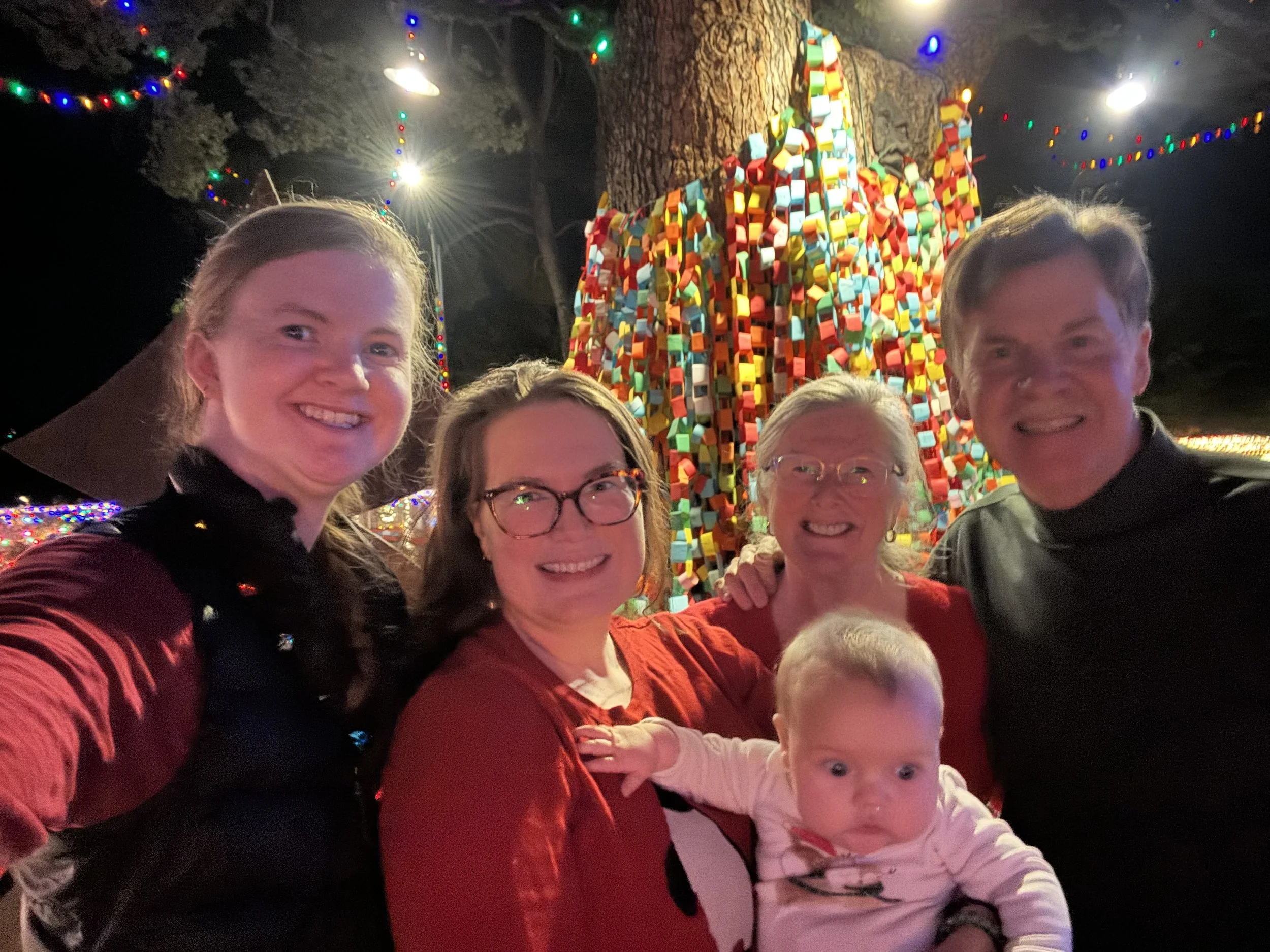 The Wishing Tree family is posing in front of a large, colorful, covered Christmas tree with lights strung in the background. Everyone is smiling.