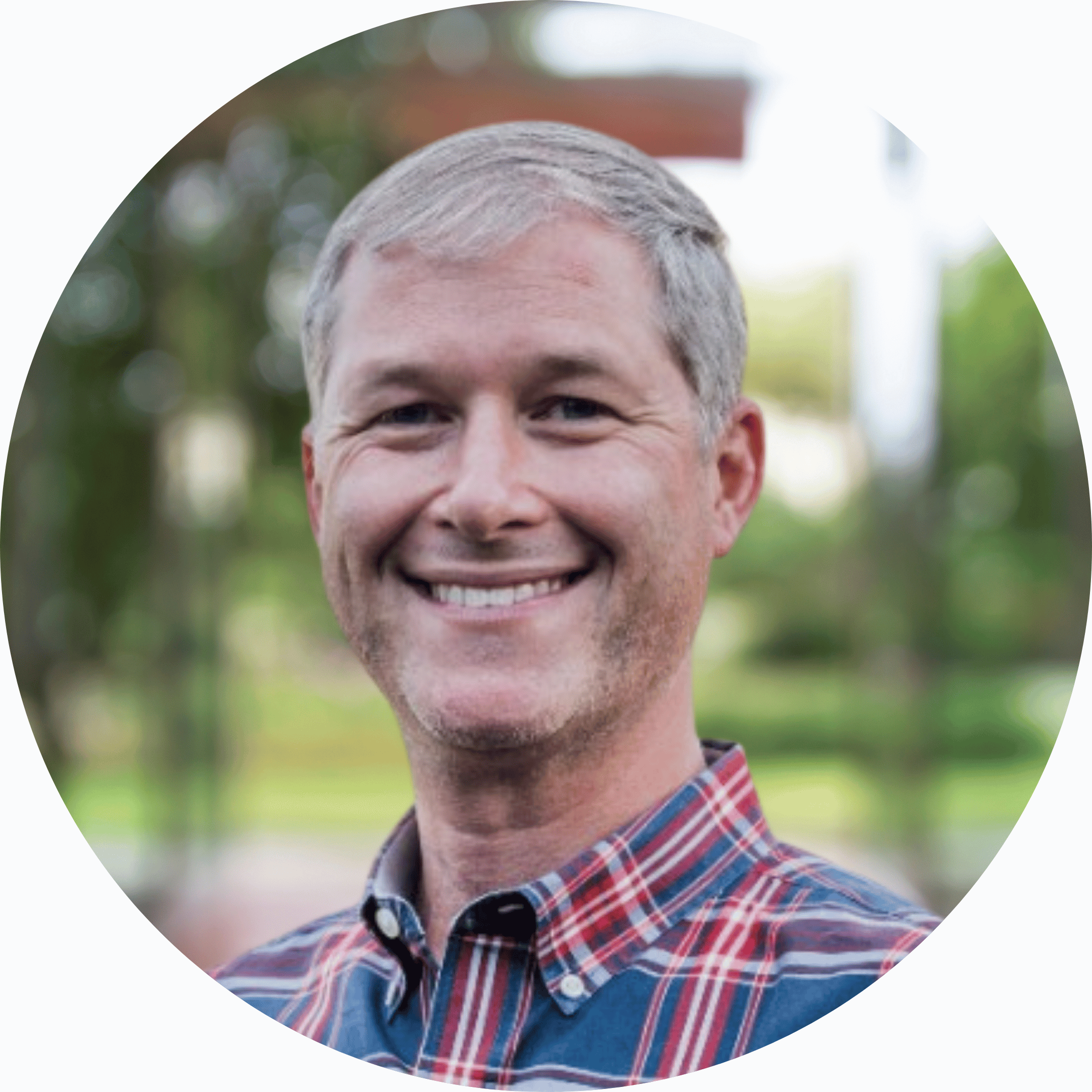 Smiling man with short gray hair wearing a plaid shirt outdoors, blurred greenery in the background.