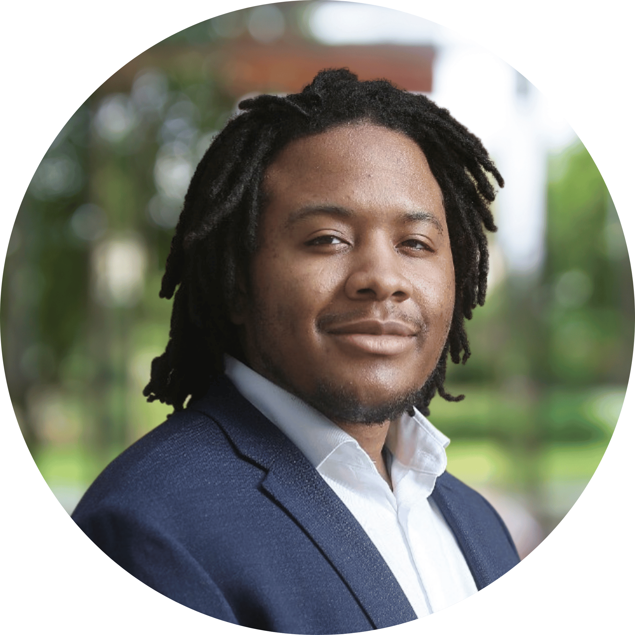 Portrait of a man with dreadlocks, wearing a navy blazer and white shirt, outdoors with blurred greenery in the background.