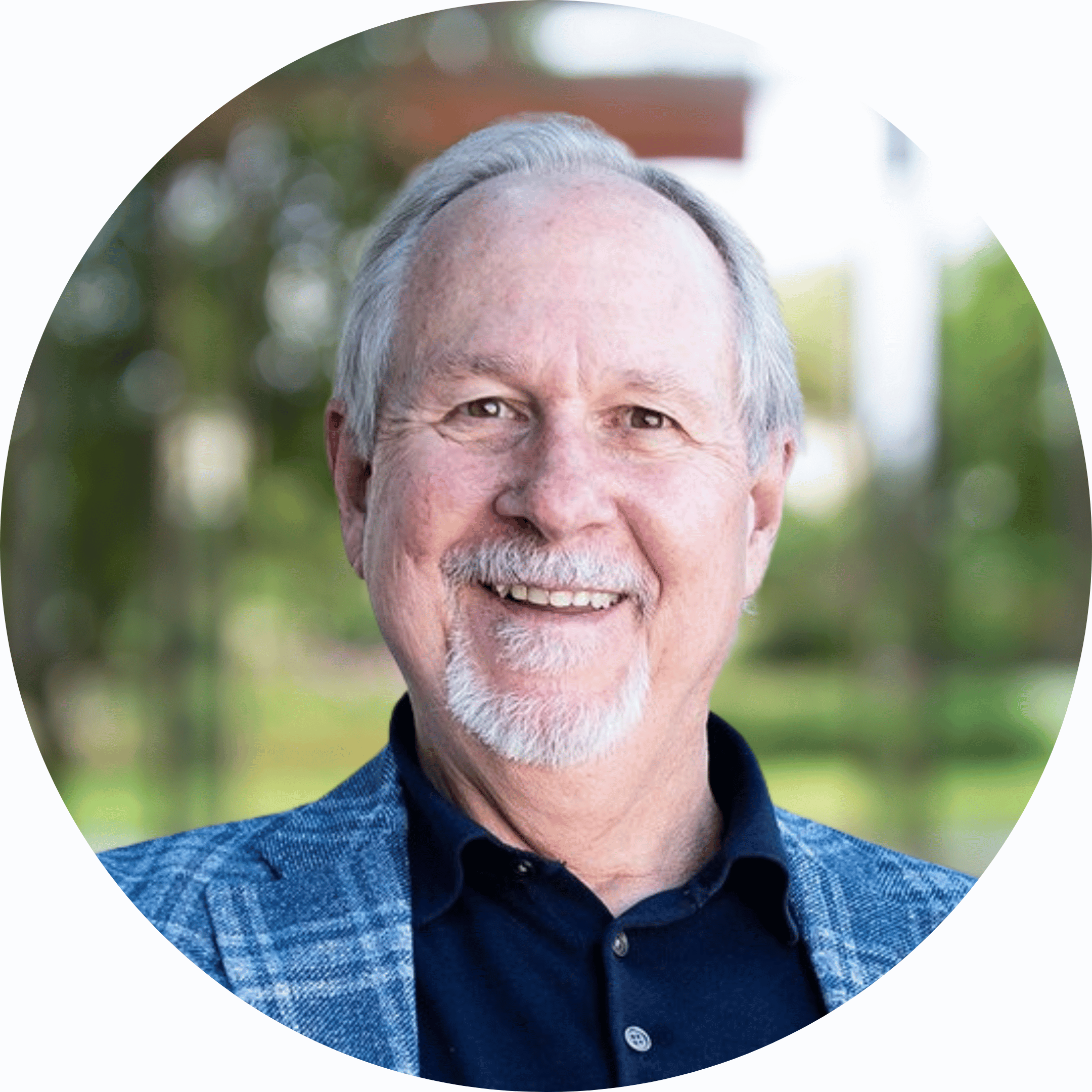 A smiling older man with gray hair, a beard, and mustache, wearing a blue patterned blazer and dark blue shirt, outdoors with blurred trees and greenery in the background.