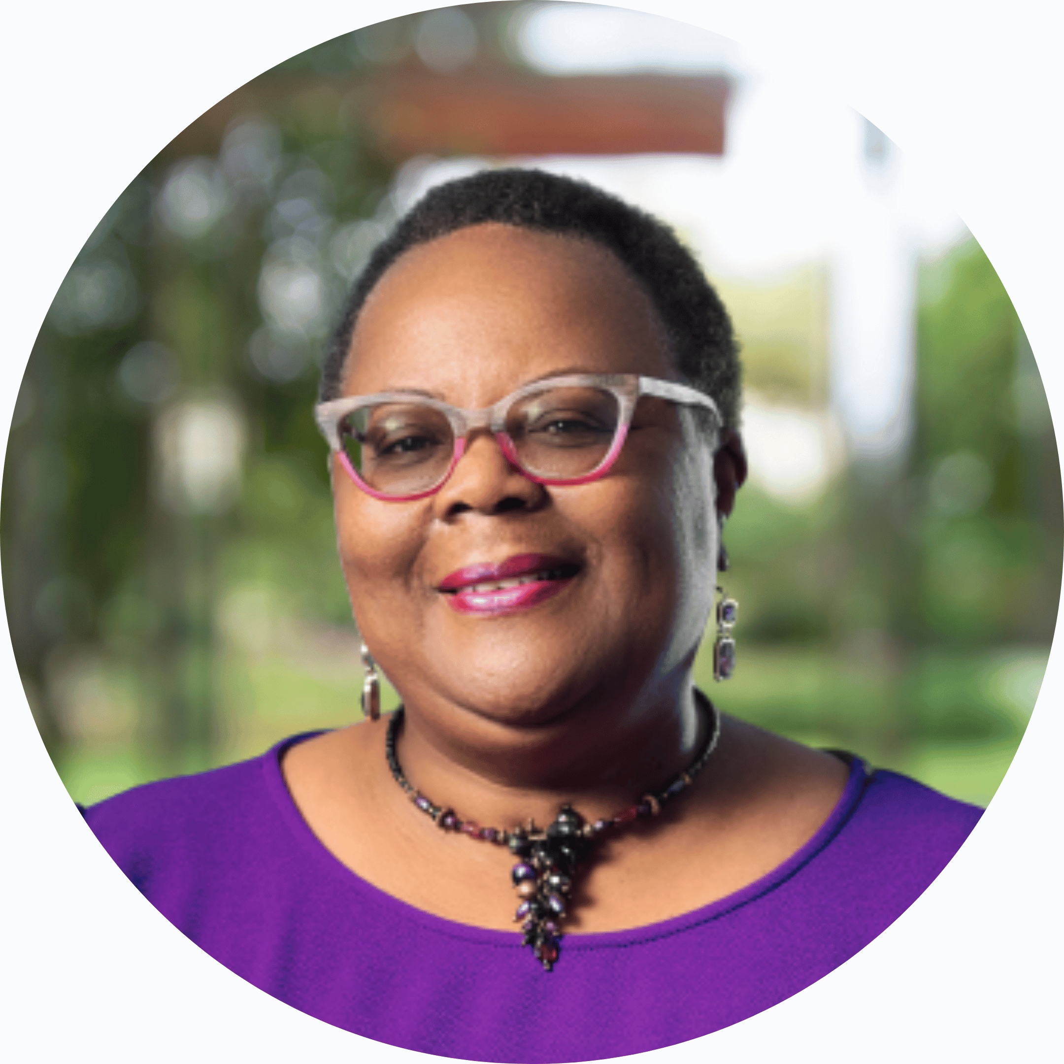 A portrait of a middle-aged African American woman with short hair, wearing colorful glasses, a purple top, and jewelry, standing outdoors with a blurred green background.