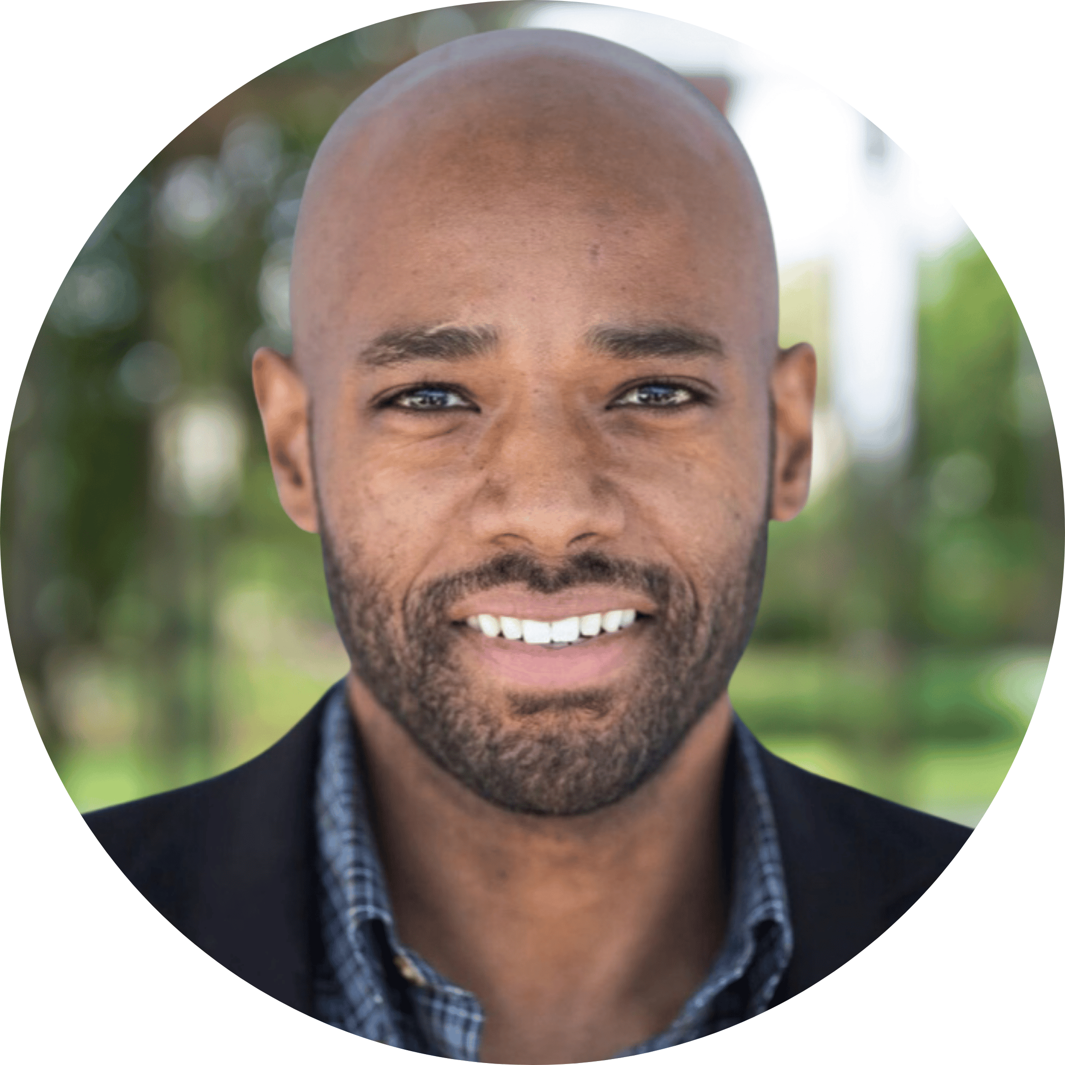 Close-up portrait of a smiling Black man with a shaved head, beard, and mustache, wearing a dark blazer and a patterned shirt, standing outdoors with a blurred green background.