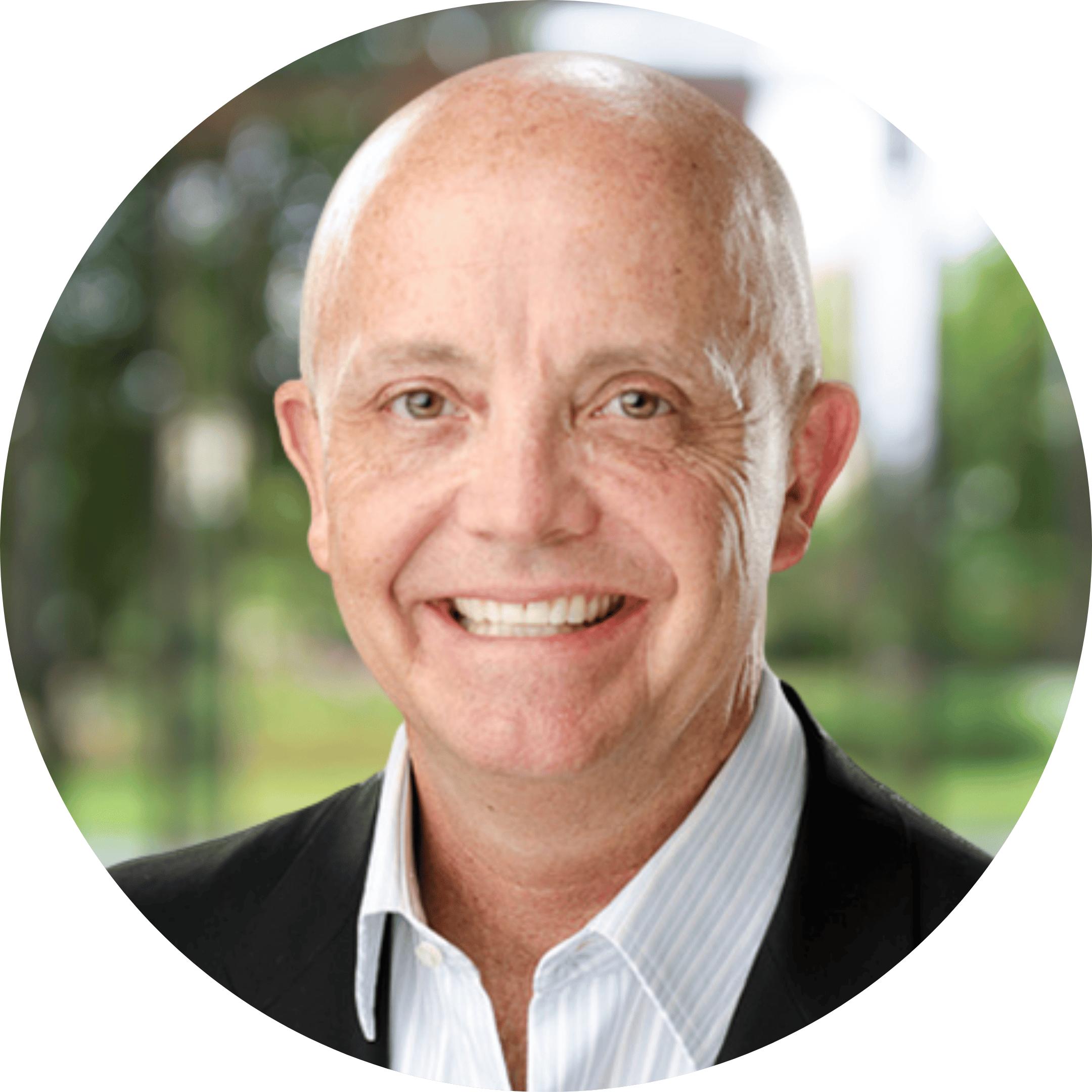 Headshot of a smiling middle-aged man with a bald head, wearing a black jacket and a white shirt, outdoors with trees and blurred background.