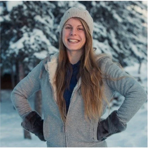 A young woman with long blond hair smiling outdoors in a snowy landscape, wearing a gray zip-up jacket, a knitted beanie, and black gloves.