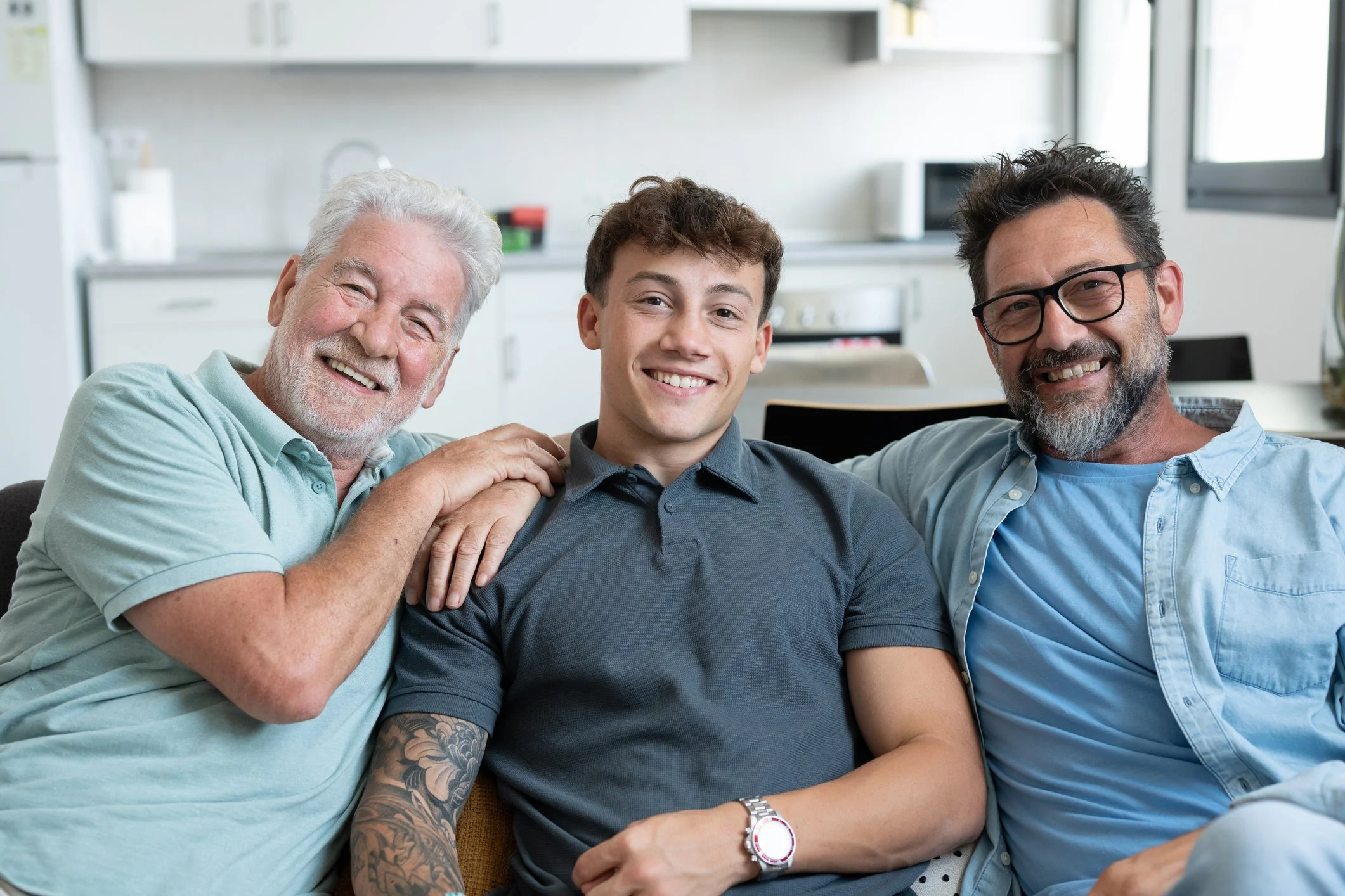 Happy multigenerational family of males stay together sitting on sofa smiling looking at camera. Grandfather, mature son and young grandson share positive moments in total relax