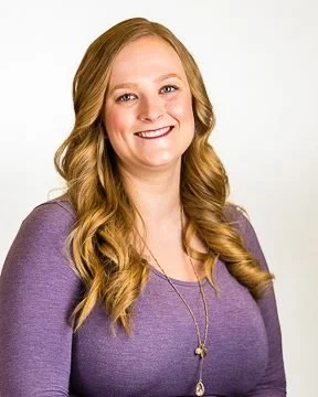 A young woman with long, wavy red hair, smiling and wearing a purple top and a necklace, posing against a white background.