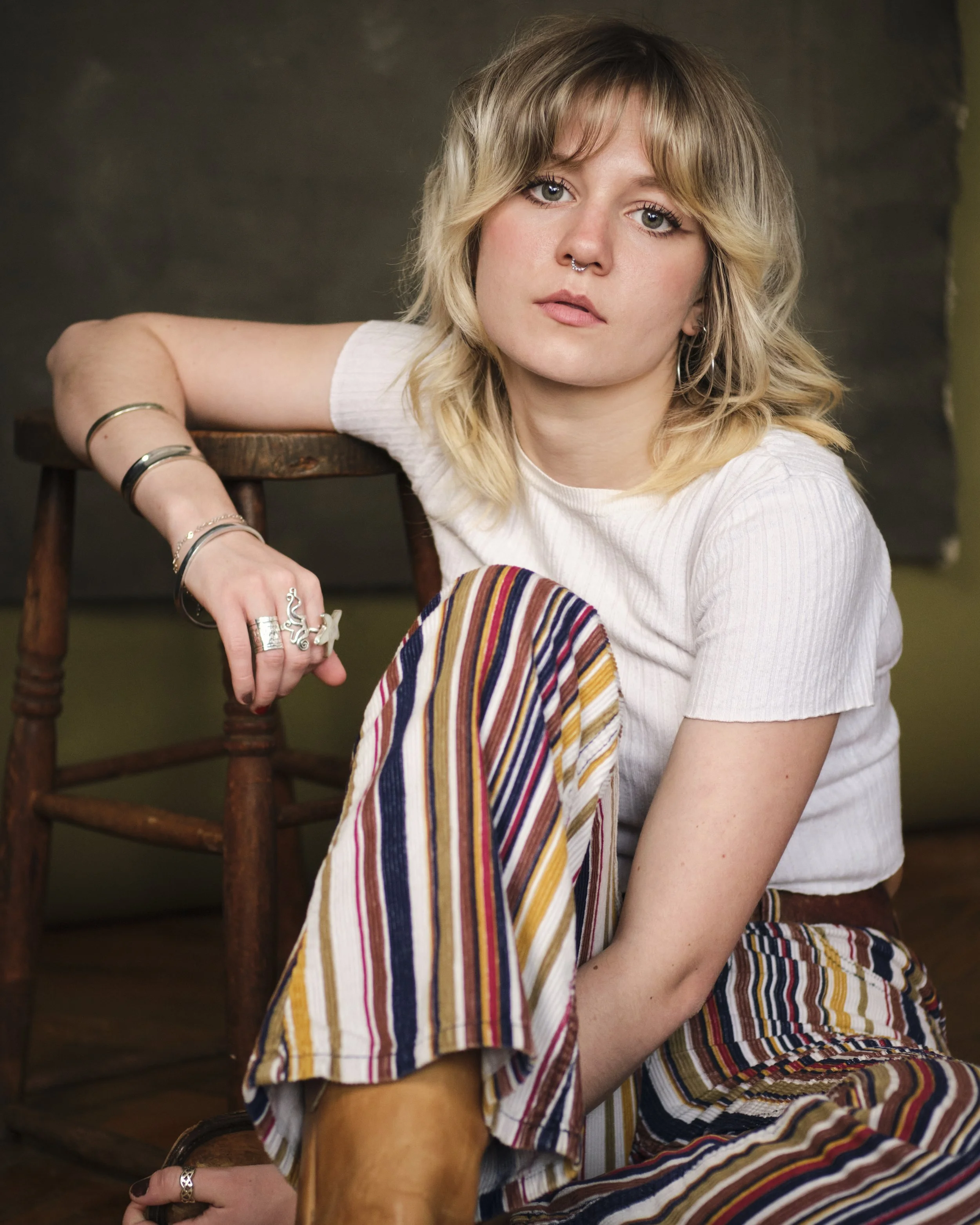 A young woman with blonde, wavy hair and light green eyes, wearing a white shirt and colorful striped pants, sitting with her arm resting on a wooden chair, against a dark, blurred background.
