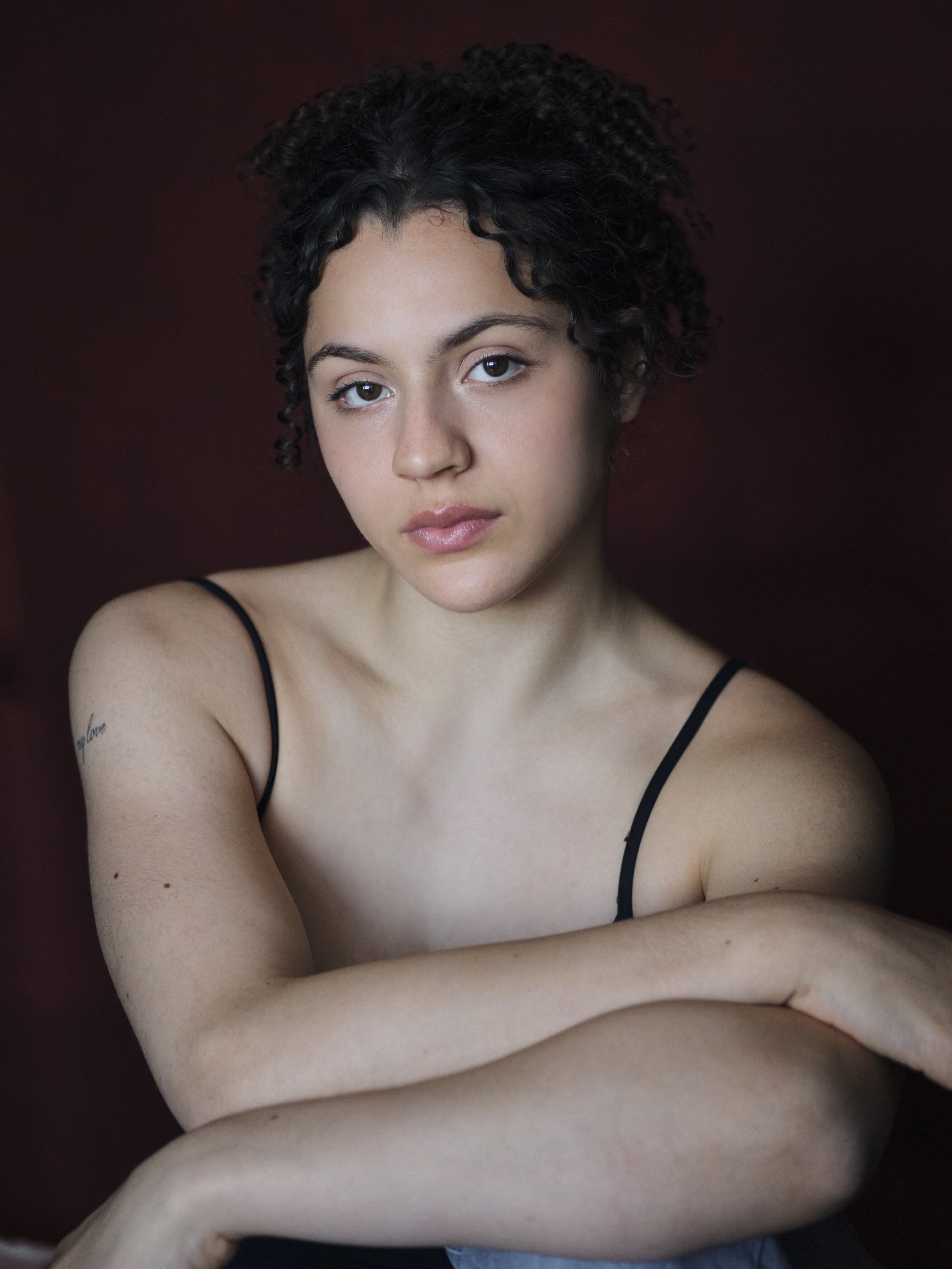 A young woman with curly dark hair looking at the camera against a dark red background. She is wearing a black spaghetti strap top, and has a tattoo on her left arm that says 'love'.