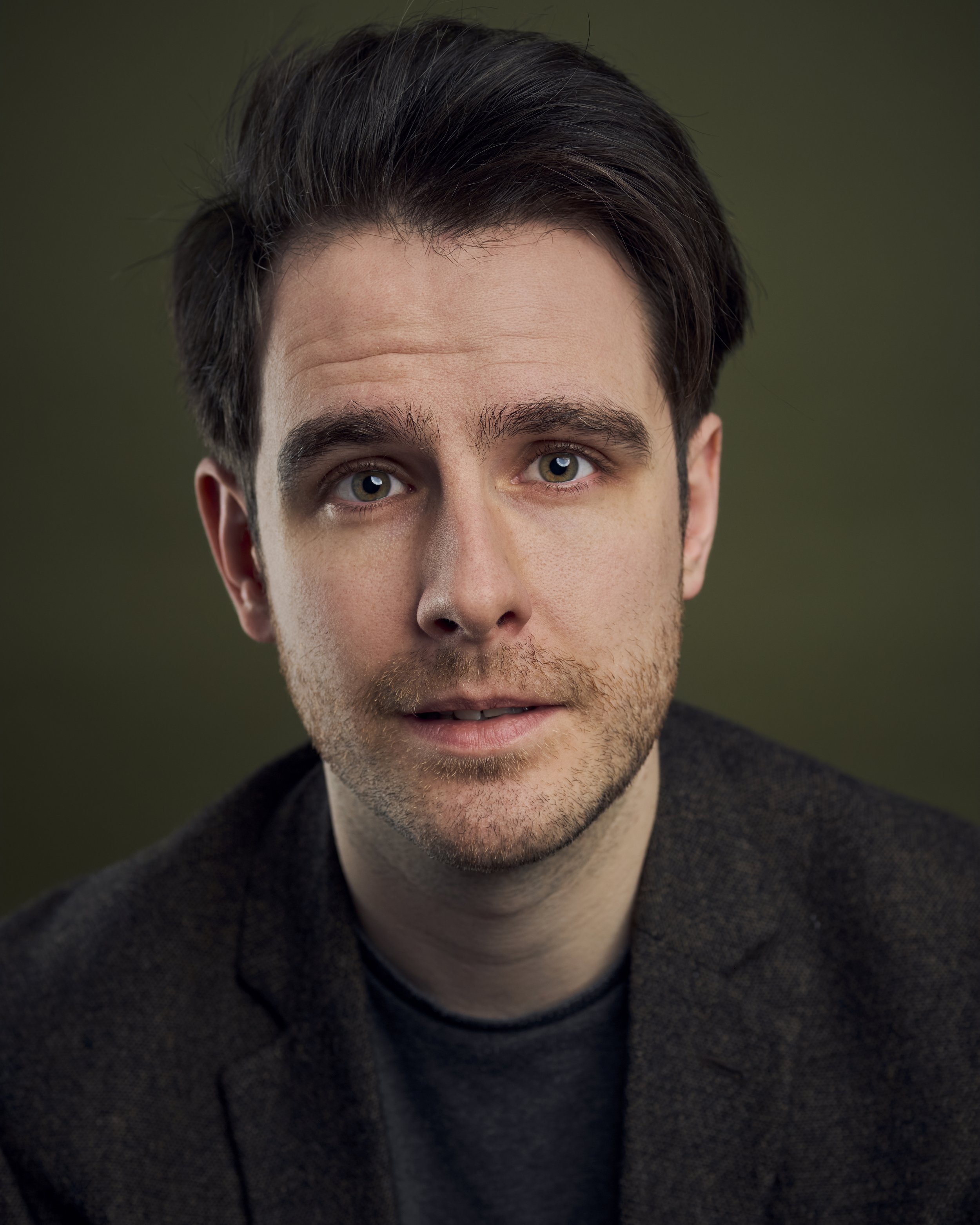 A close-up portrait of a young man with dark hair, light skin, and hazel eyes, wearing a dark sweater against a dark green background.