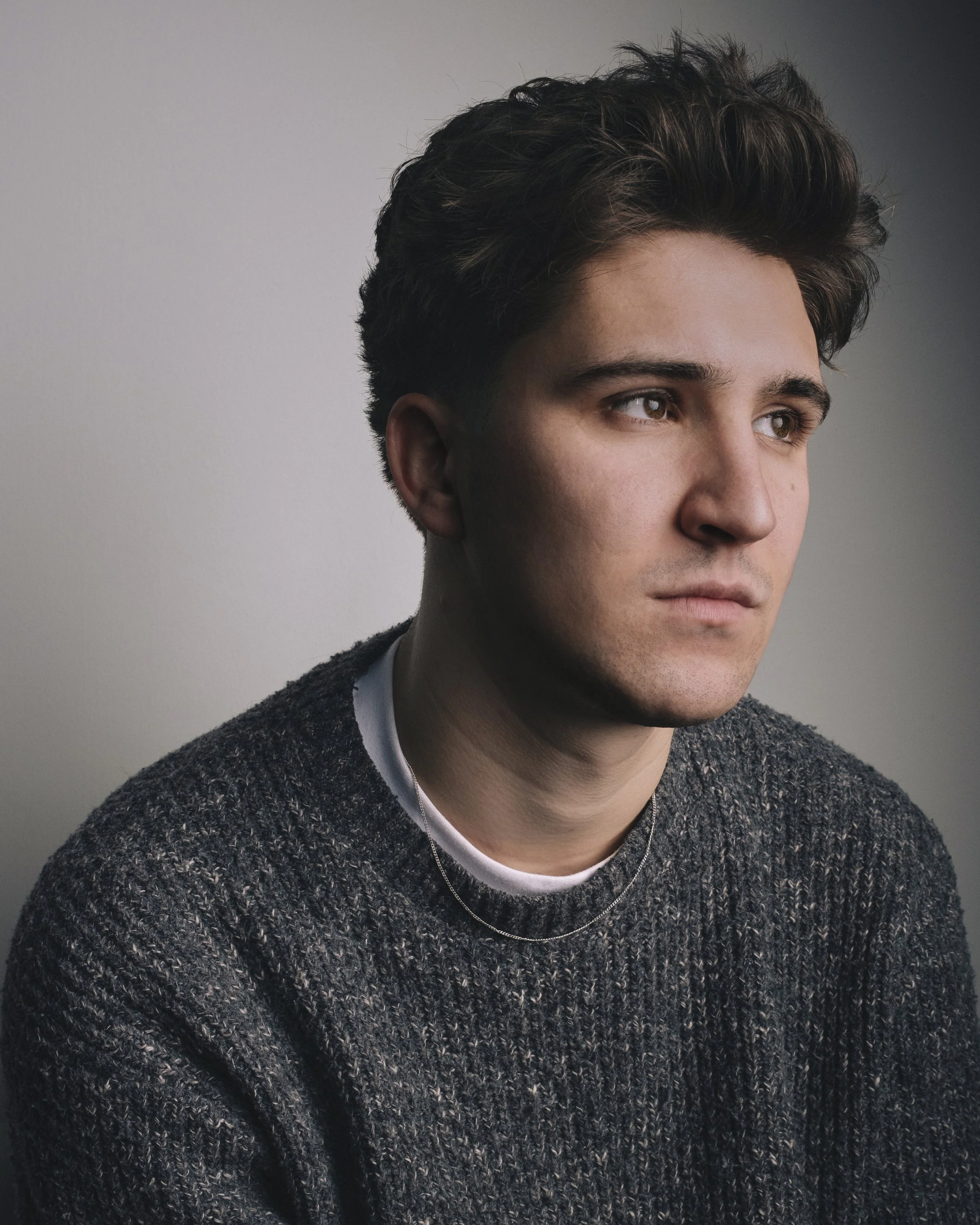 Close-up of a young man with brown eyes, dark wavy hair, wearing a dark textured sweater, layered necklaces, and a grey shirt, against a plain grey background.