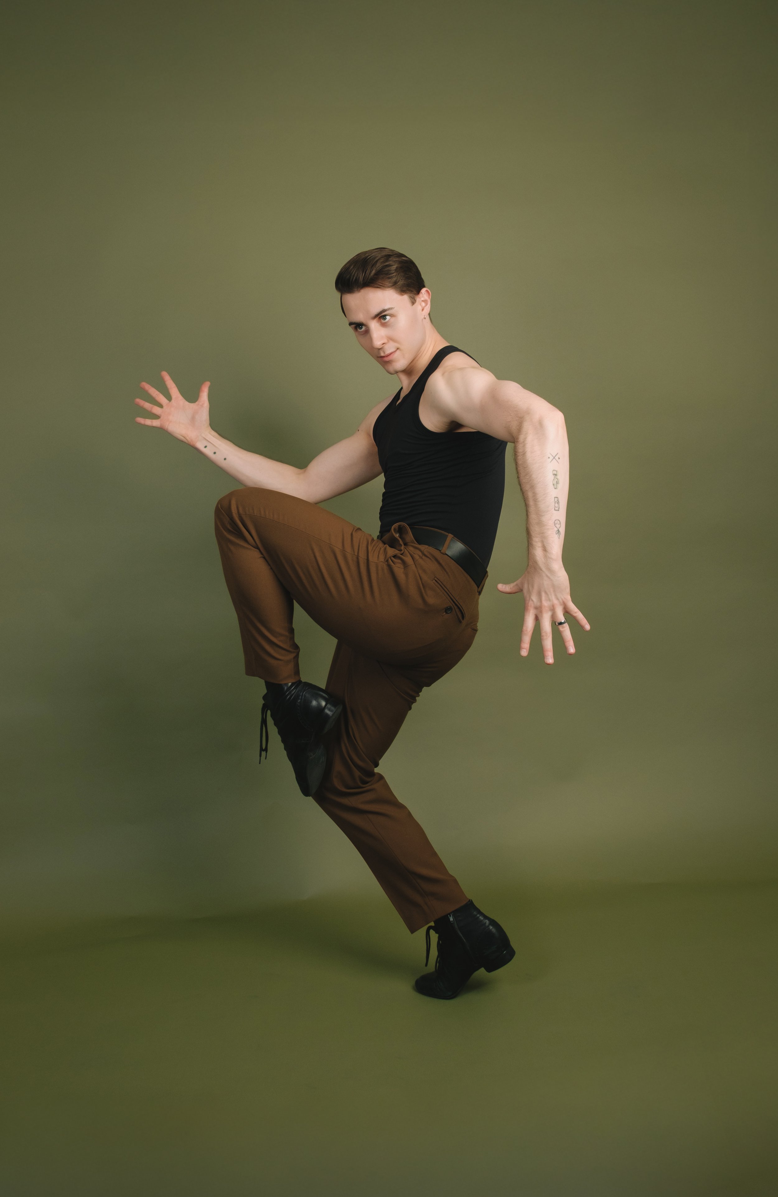 A person with dark, short hair and tattoos on their arms poses against a plain green background. They are wearing a black tank top, brown pants, and black boots, balancing on one foot with their other knee raised and arms extended outward.
