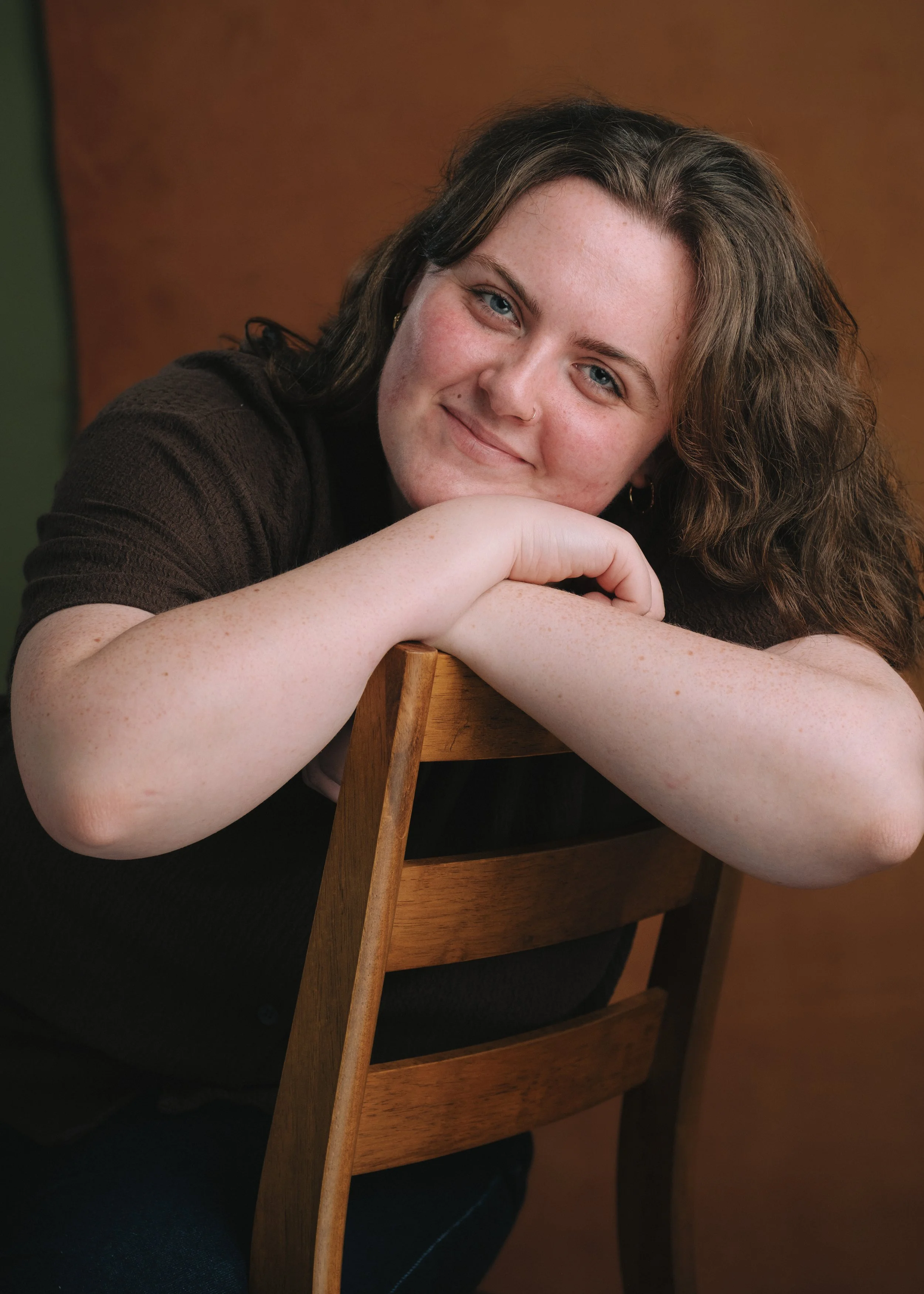 A woman with brown hair and blue eyes smiling, resting her head and arms on a wooden chair back, wearing a black top, with a plain brown background.