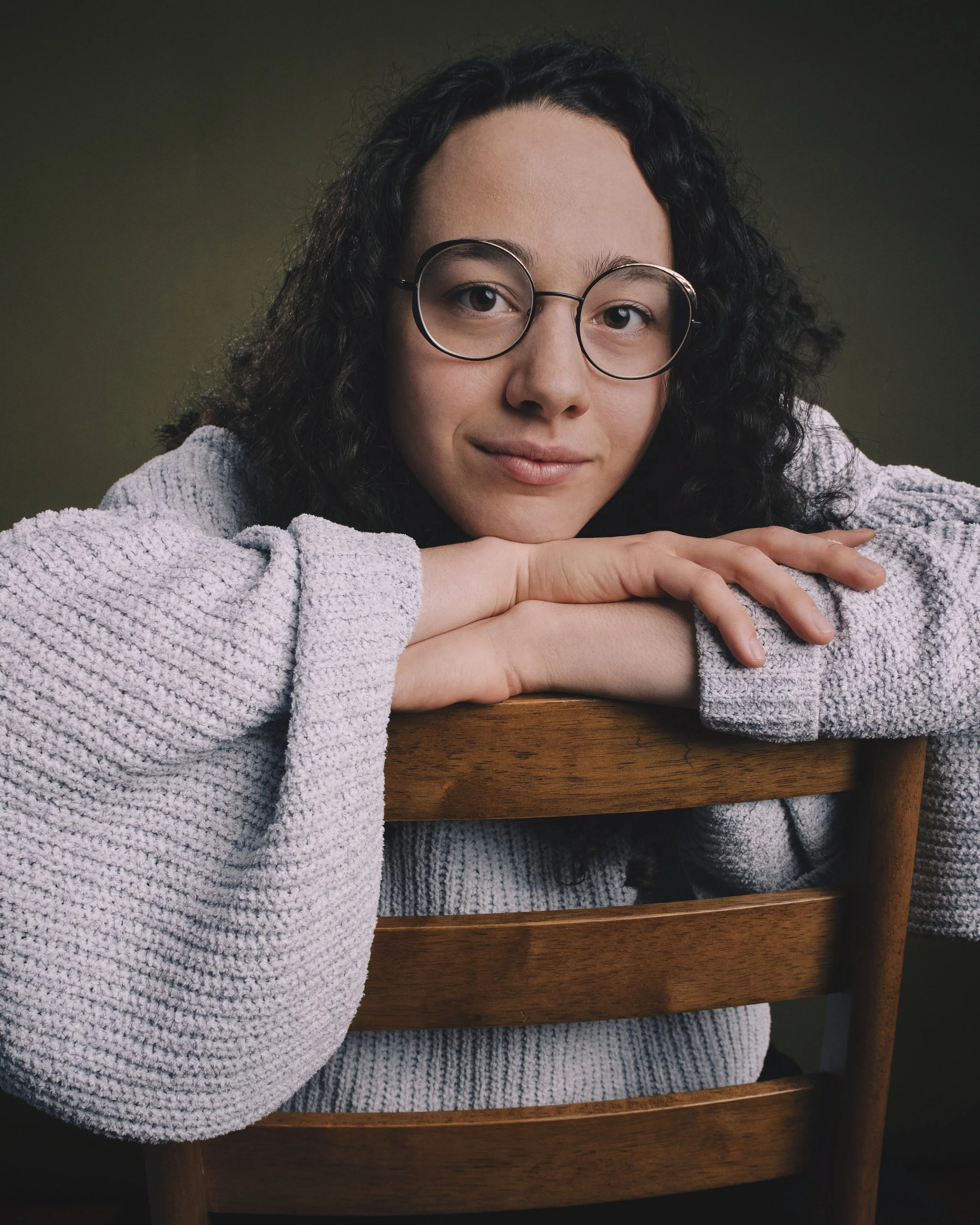 A woman with curly dark hair and glasses, wearing a fluffy grey sweater, resting her chin on her crossed arms on a wooden table, with a neutral background.