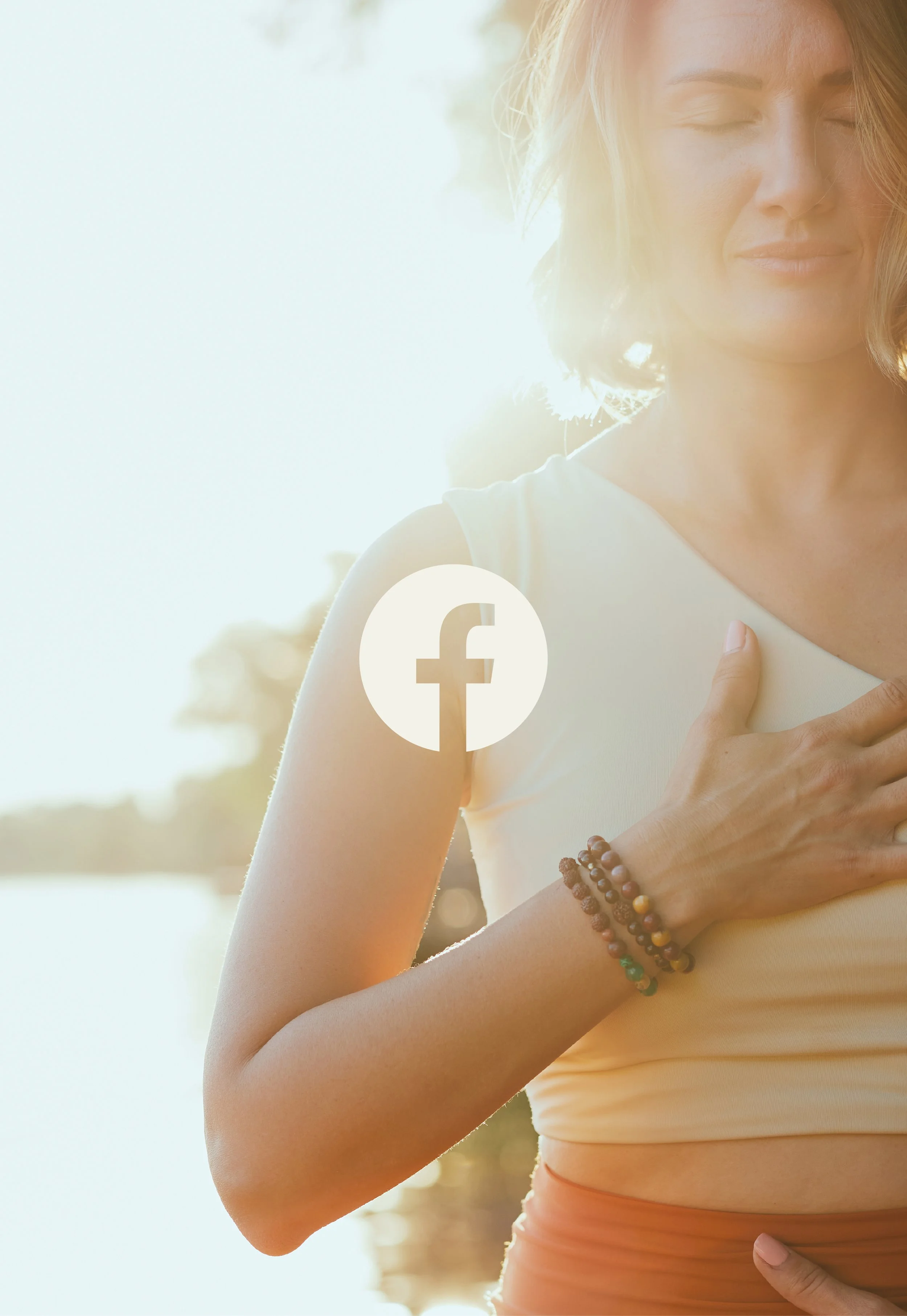 A woman with short blonde hair outdoors with her eyes closed, gently placing her hand on her chest and another on her stomach, wearing a white sleeveless top and beaded bracelets on her wrist, with sunlight in the background.
